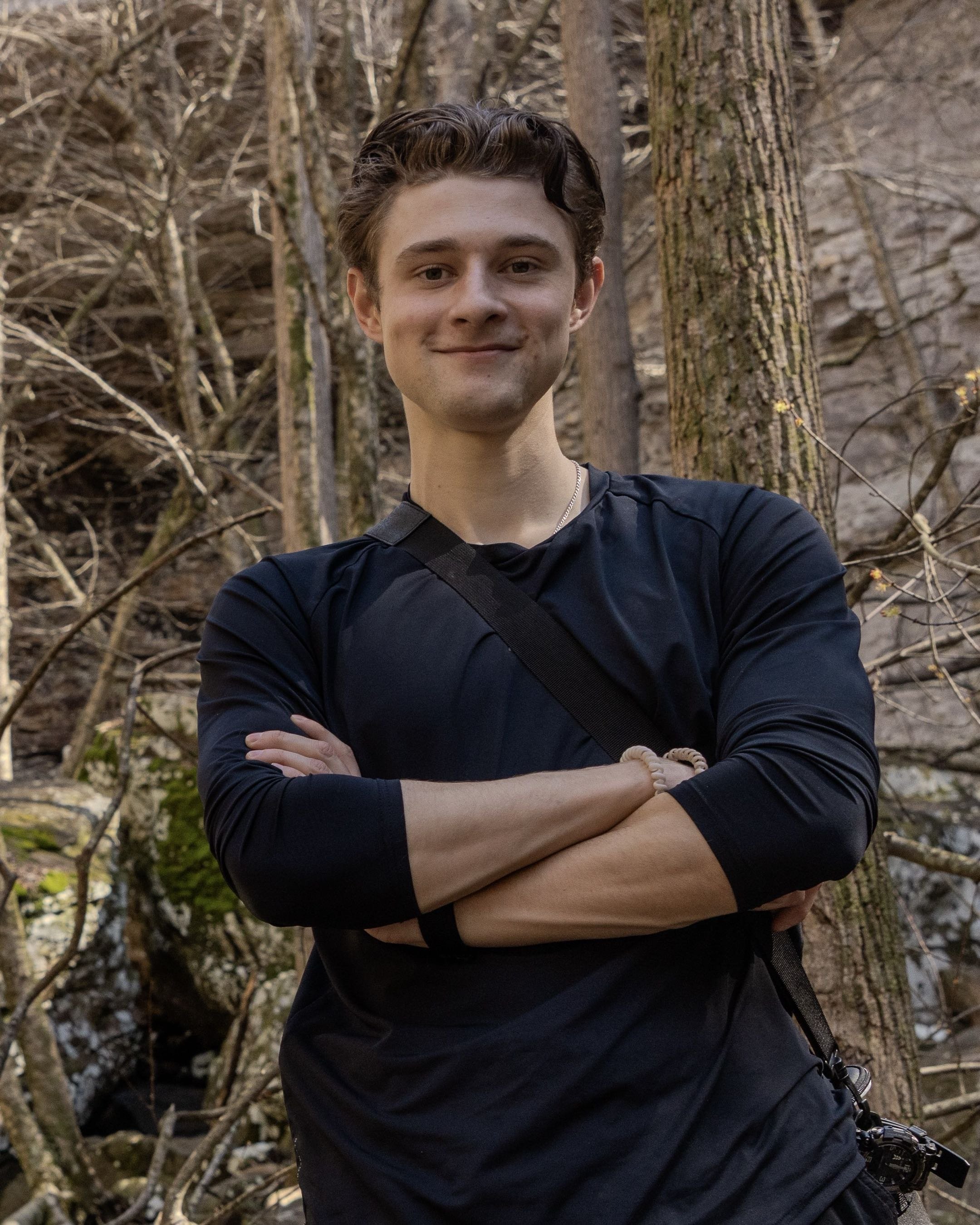 A young man with light skin and wavy brown hair, smiling with arms crossed in front of a forested background with trees and rocks.