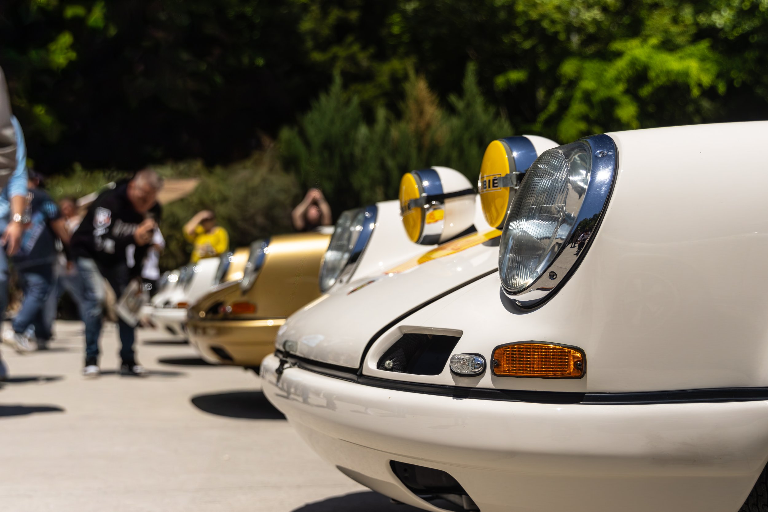 Close-up of white vintage cars parked in a row during the daytime with people in the background, trees, and clear sky.