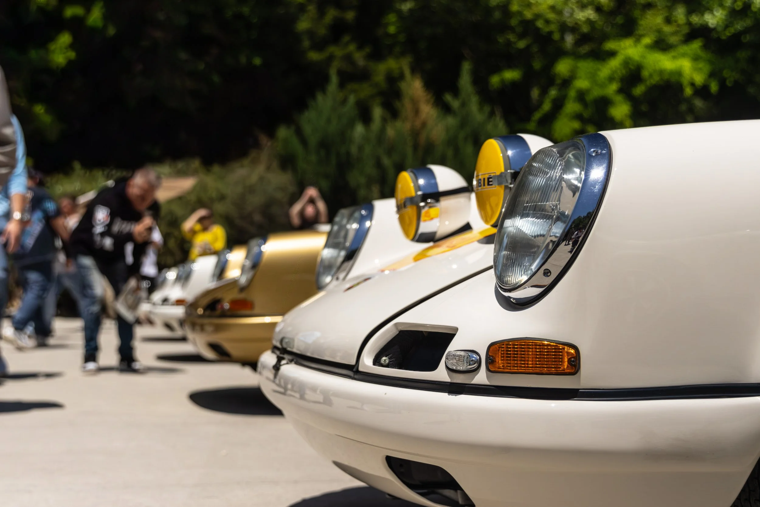 Close-up of the front of a white vintage car, showing its headlight, turn signal, and part of the bumper. People are gathered in the background, some taking photos, with trees and sunlight in the outdoor setting.