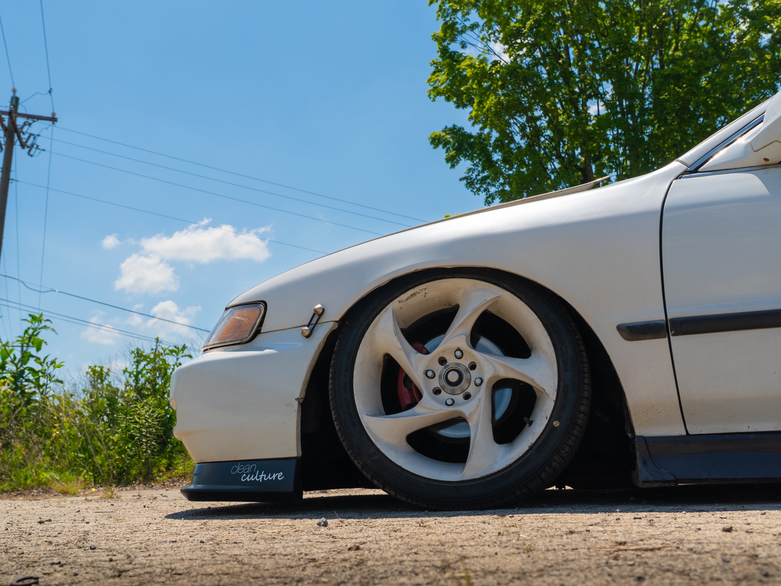 Close-up of a white sports car parked outdoors under a clear blue sky with green trees in the background.