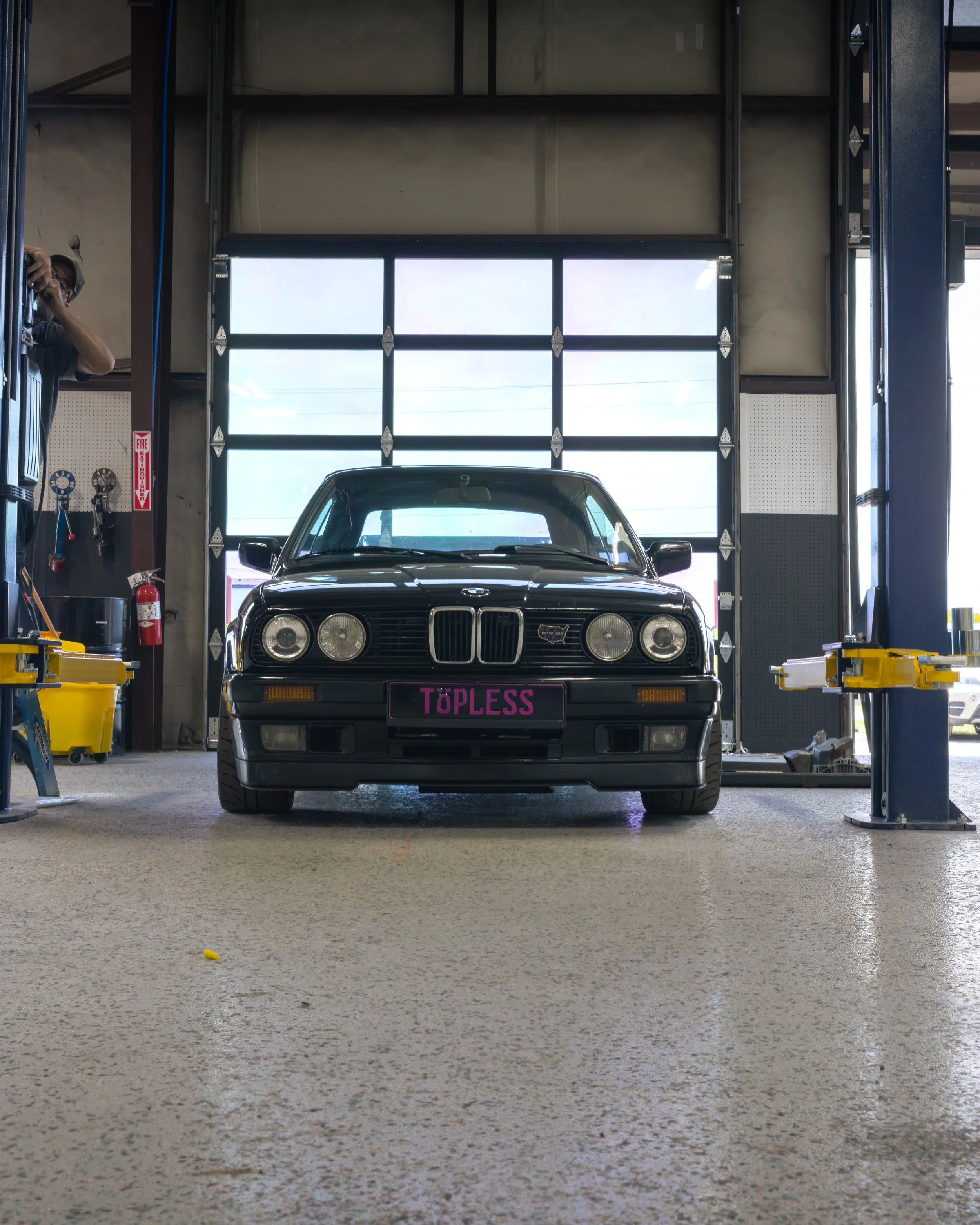 A black BMW car inside a garage with a pink custom license plate reading 'TØPLESS'.