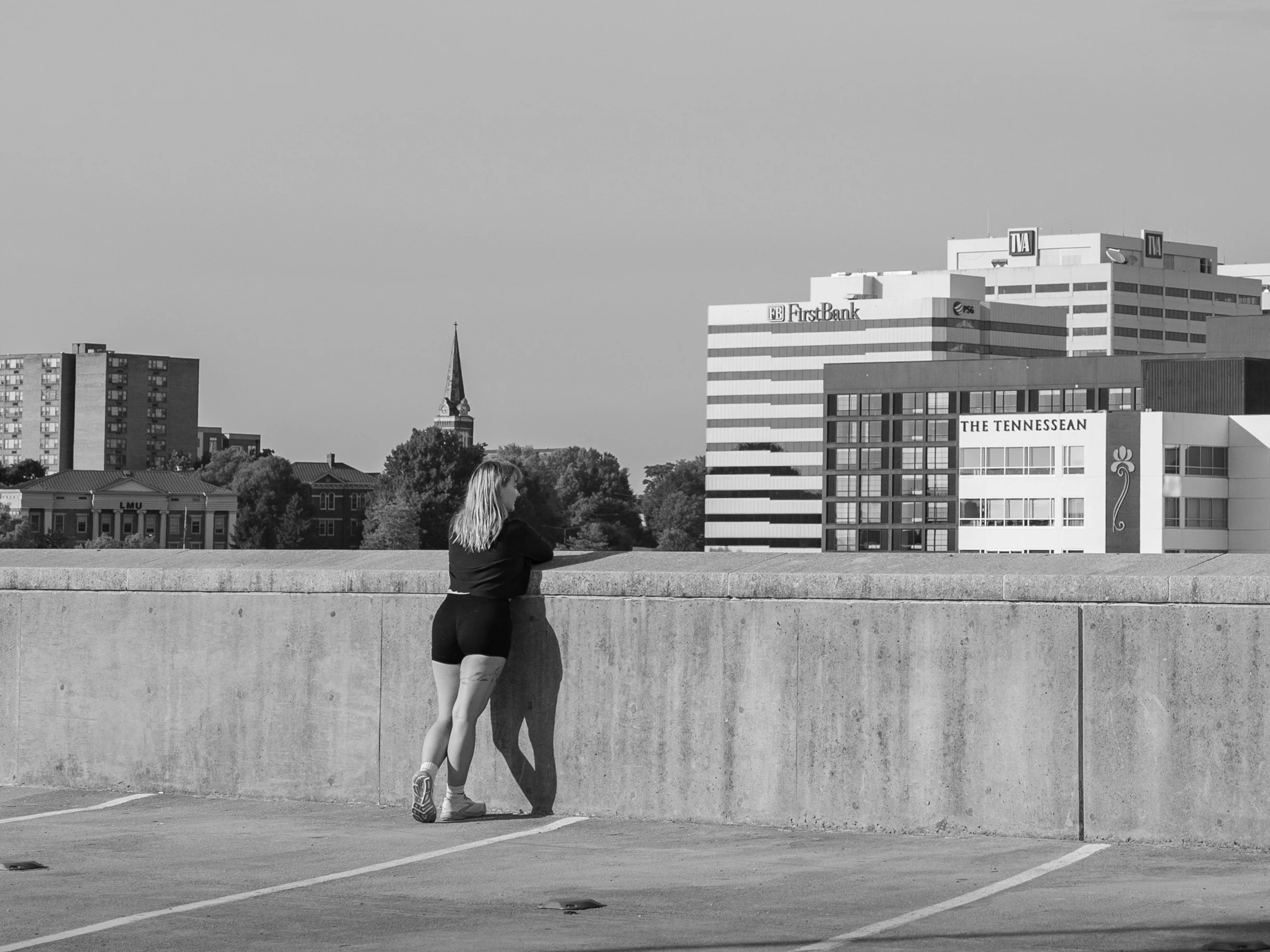 A woman in athletic clothing leaning against a concrete wall in a parking lot with city buildings in the background, including a church steeple and several commercial office buildings, some with signs like FirstBank and TVA, in black and white.