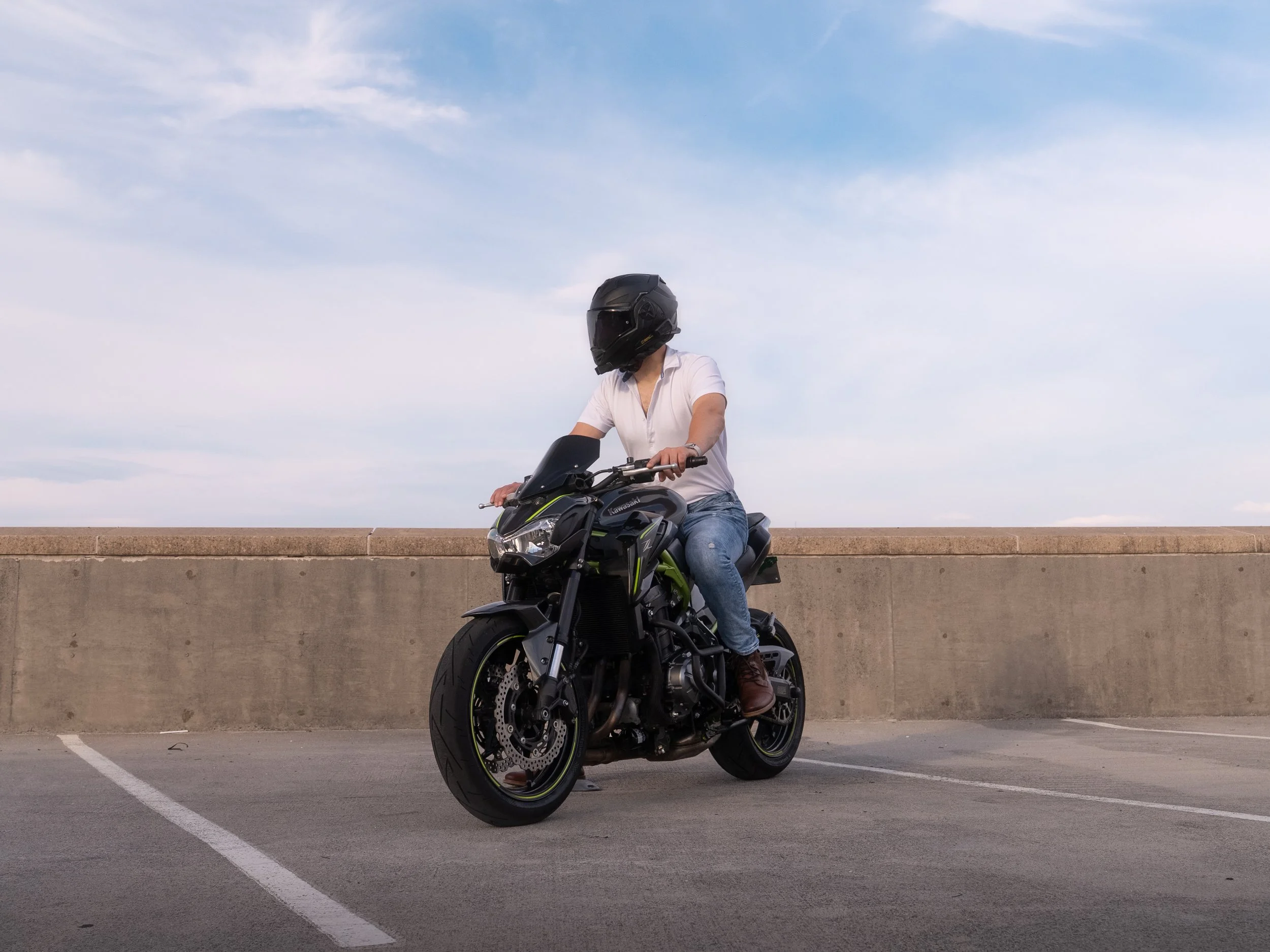 A person riding a black and green Kawasaki motorcycle in a parking lot, with a concrete barrier and blue sky with some cloud streaks in the background.