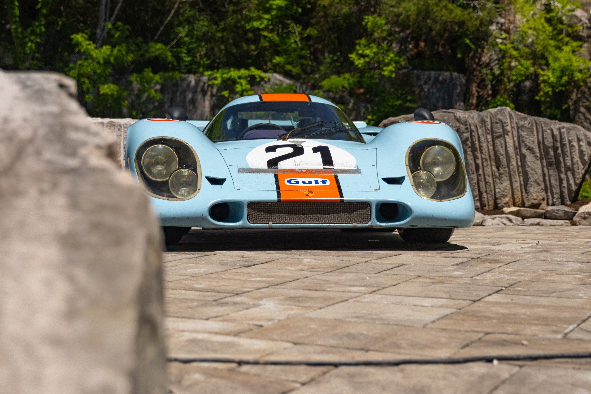 A vintage blue racing car with the number 21 on the front, partially obscured by a large rock in the foreground, parked outdoors with green foliage in the background.