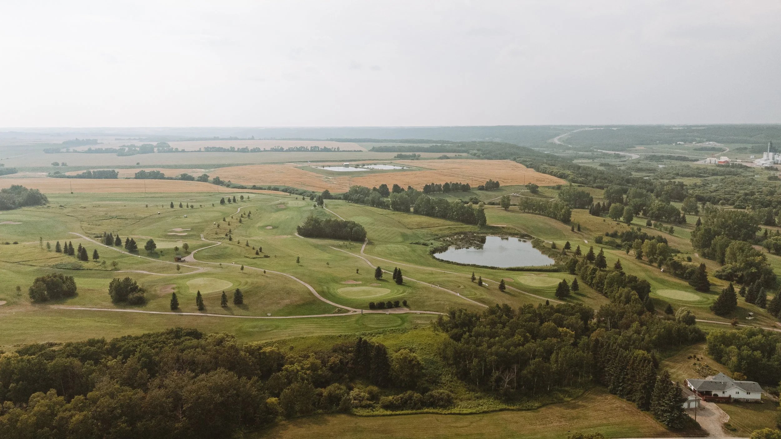 Aerial view of a golf course with green fairways, water hazards, trees, and surrounding farmland in the distance under a cloudy sky.