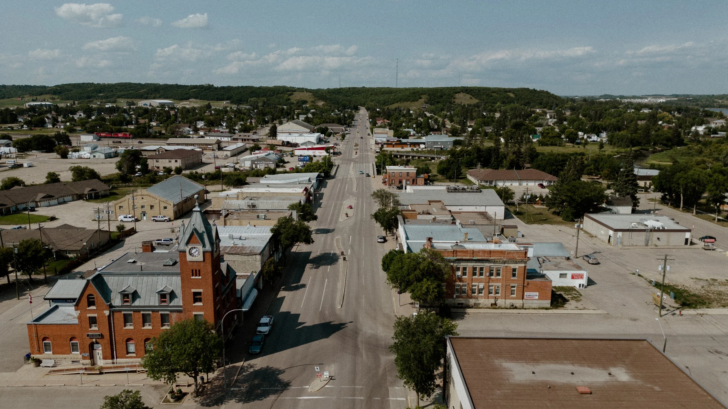 Aerial view of a small town with a main street, historic brick buildings including a clock tower, surrounding residential areas, trees, hills in the background, and partly cloudy sky.