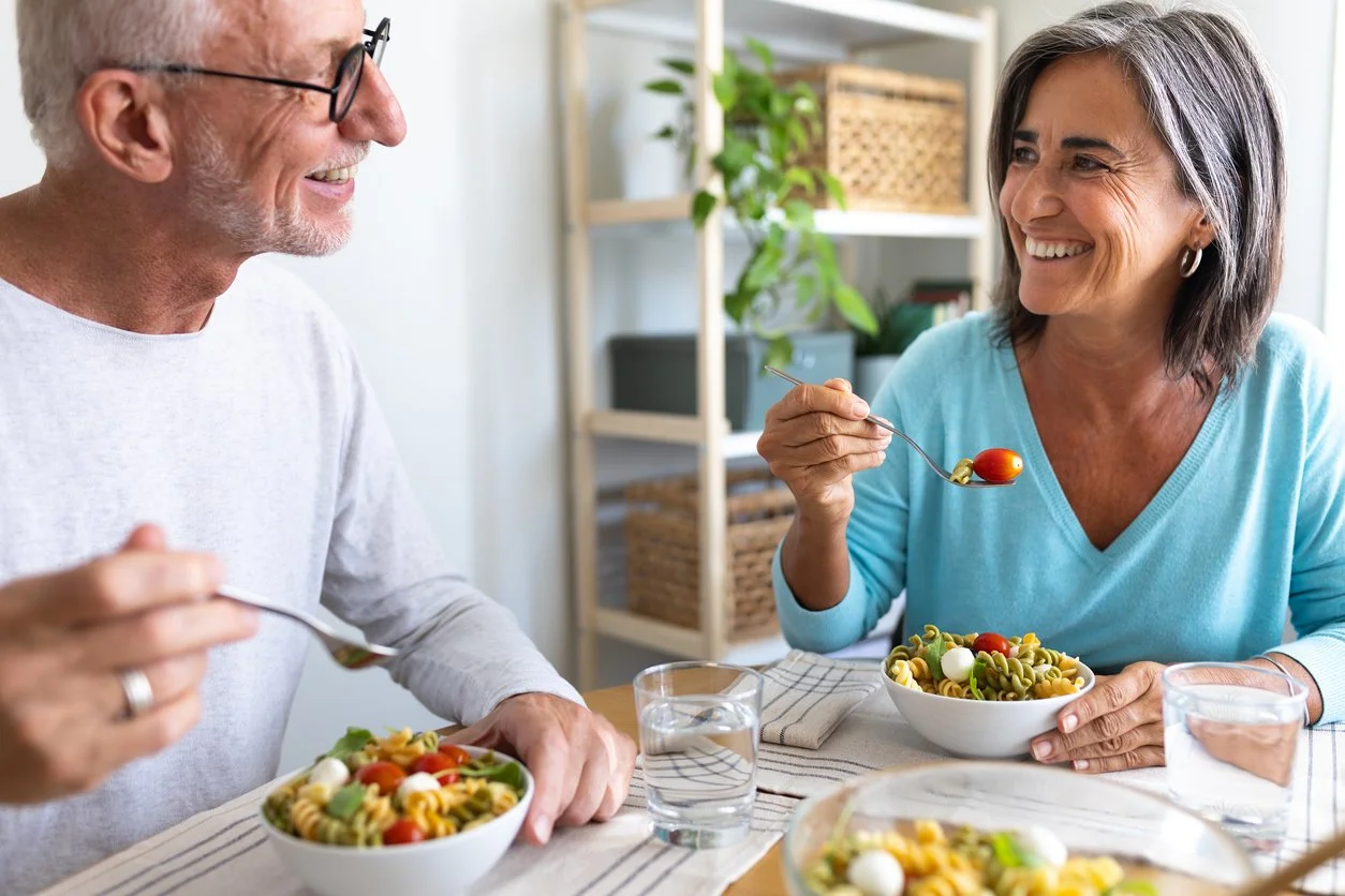 An elderly man and woman smiling and enjoying a bowl of pasta salad at a table in a bright, cozy kitchen.