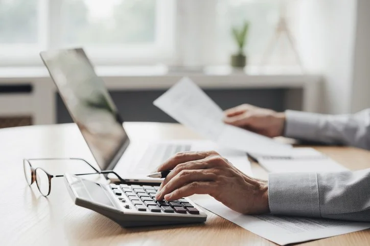 Person working at a desk with a laptop, calculator, papers, and eyeglasses.