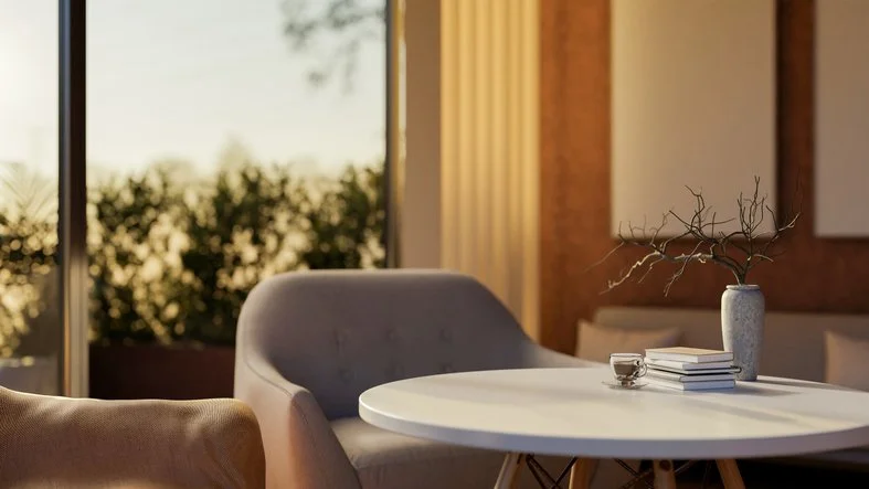 A modern dining room with a round white table, a gray upholstered chair, and a beige armchair. The table has a stack of books, a glass of water, and a tall vase with decorative branches. Large windows behind show an outdoor view of trees and a sunset.