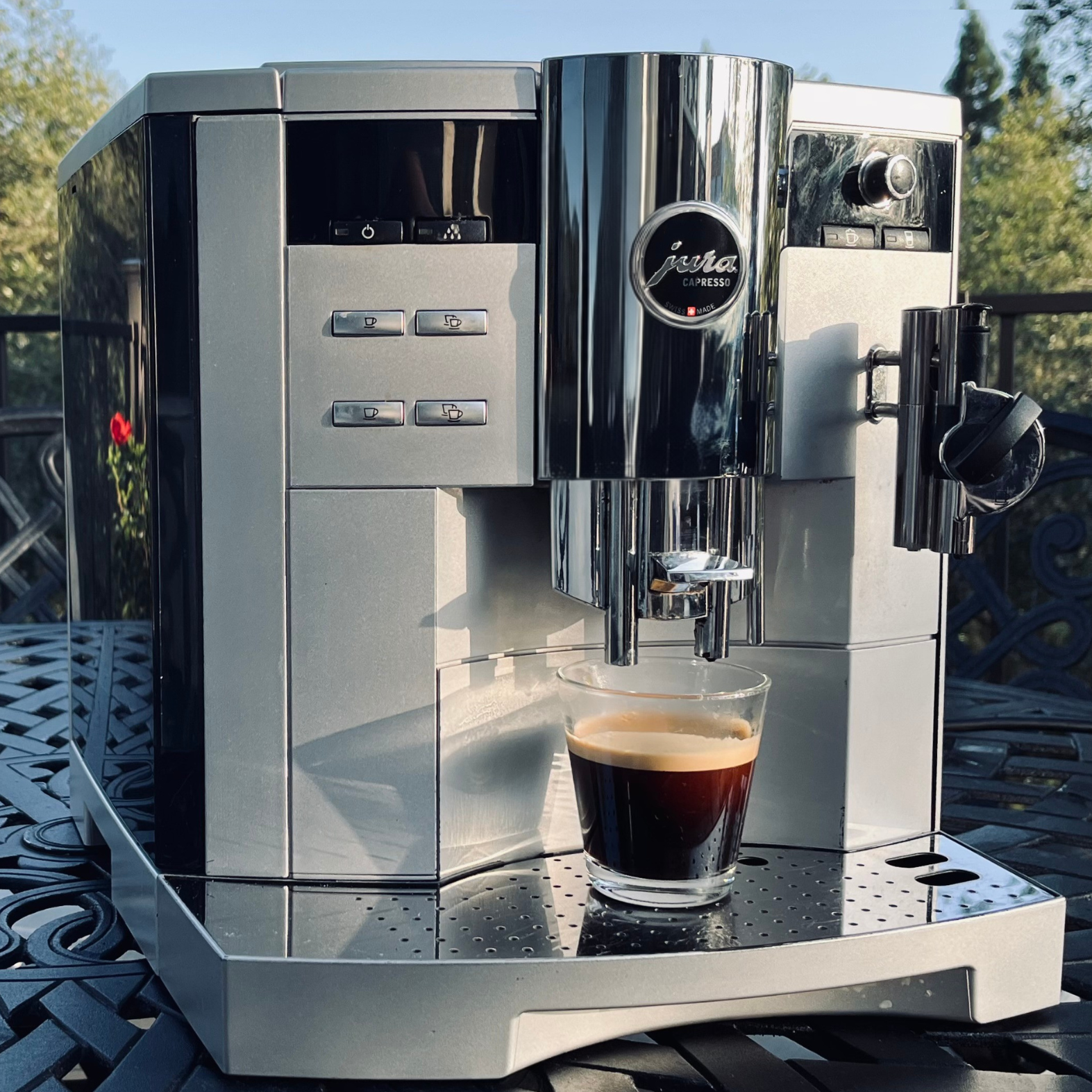 A Jura espresso machine brewing coffee into a glass cup on a metal tray, outdoors with trees and a blue sky in the background.