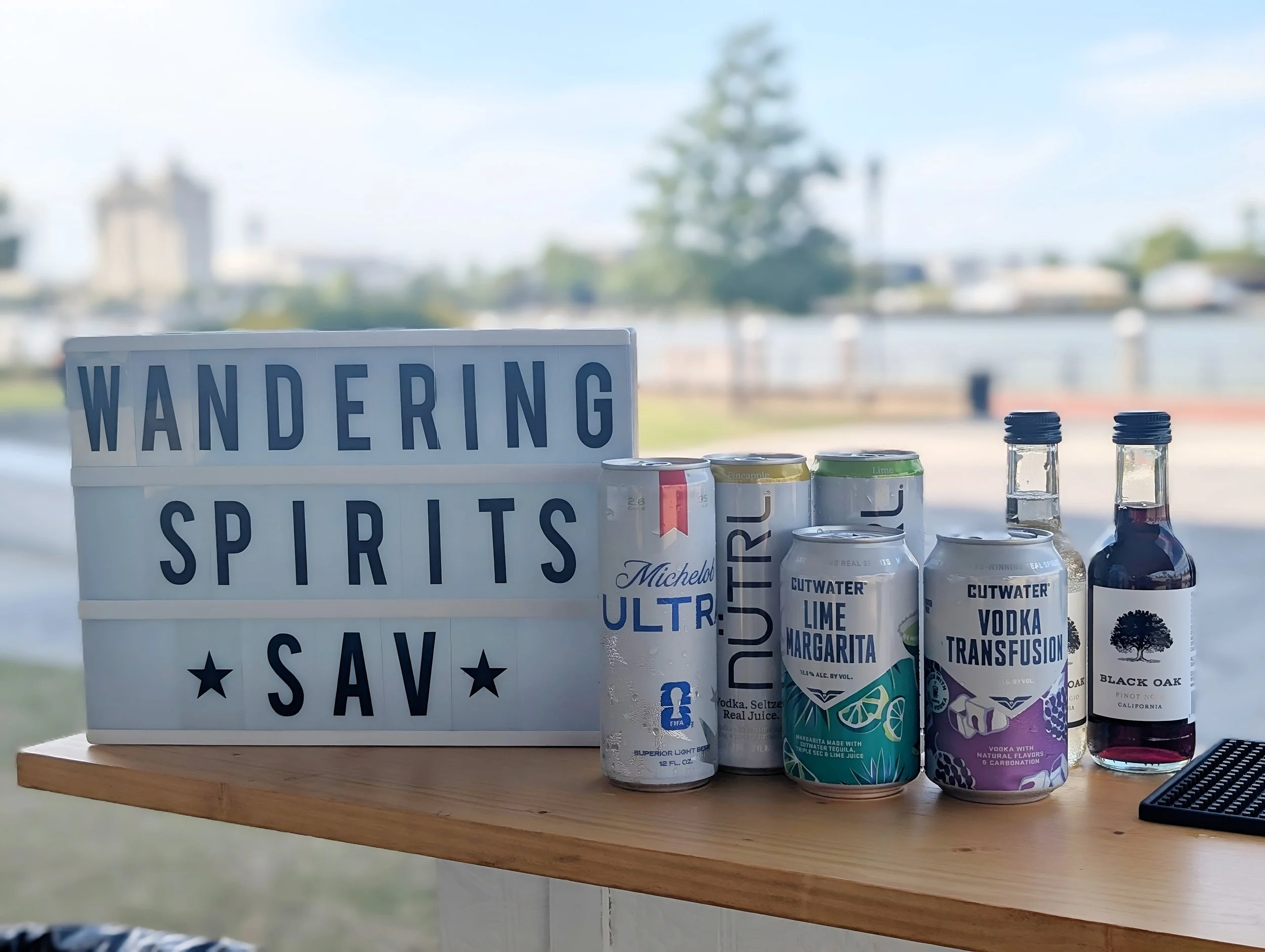 A table displays cans of alcohol and mixers with a sign that says 'Wandering Spirits, Save, Say'.