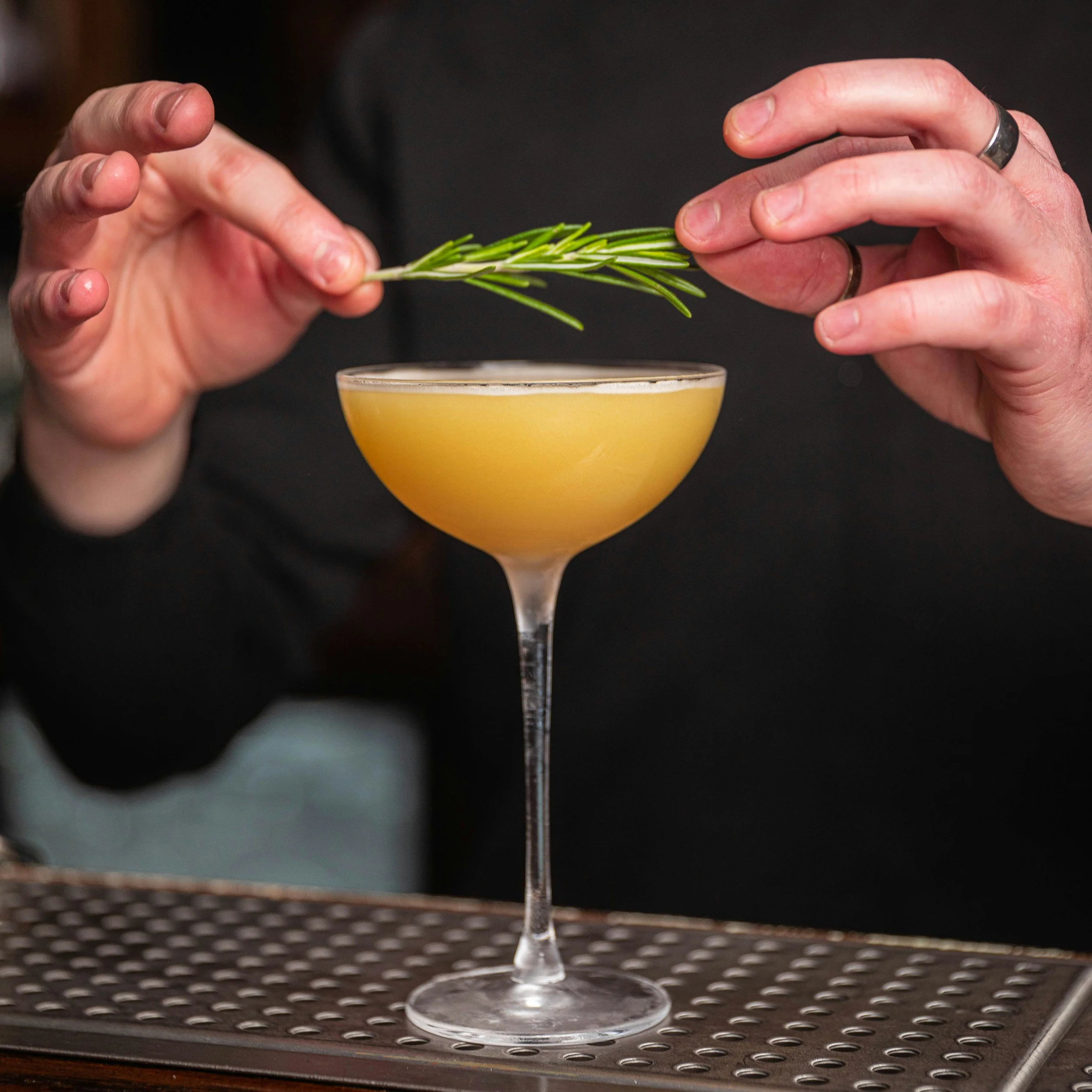 A barista's hands placing a sprig of rosemary into a yellow cocktail glass, with a textured metal surface below and a dark background.