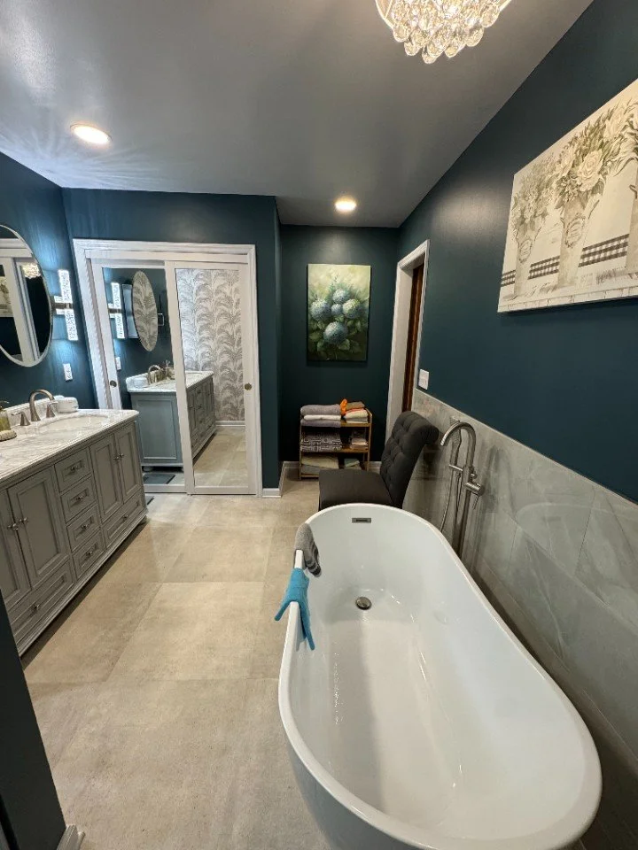 Bathroom with a standalone bathtub, a vanity with two sinks, a mirror, a chair, and wall art.