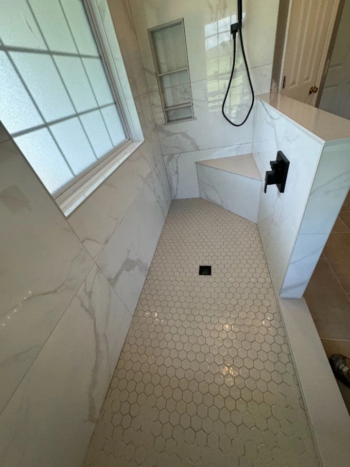 View of a modern, marble-tiled shower with hexagonal floor tiles, a built-in corner seat, a wall-mounted black shower control, a handheld showerhead, and a window with frosted glass.