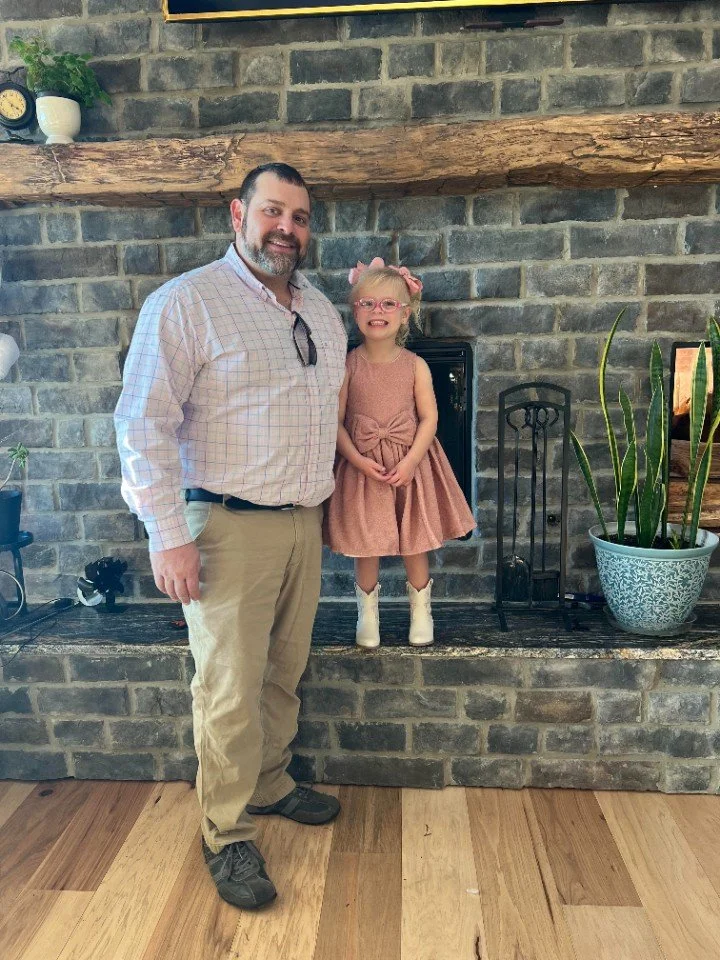 A man and a young girl standing indoors in front of a brick fireplace with a wooden mantel. The man has short dark hair and a beard, wearing a light-colored checkered shirt and khakis. The girl, standing on the fireplace ledge, is wearing glasses, a pink dress, and white boots with pink bows, smiling at the camera.