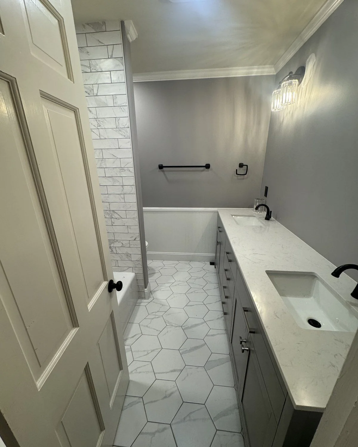 Bathroom with white hexagon tile floor, gray cabinets, white marble countertop with two sinks, black faucets, in-wall light fixture, gray painted walls, towel bar, toilet paper holder, towel hook, partially open off-white door, shower with marble and tile walls.