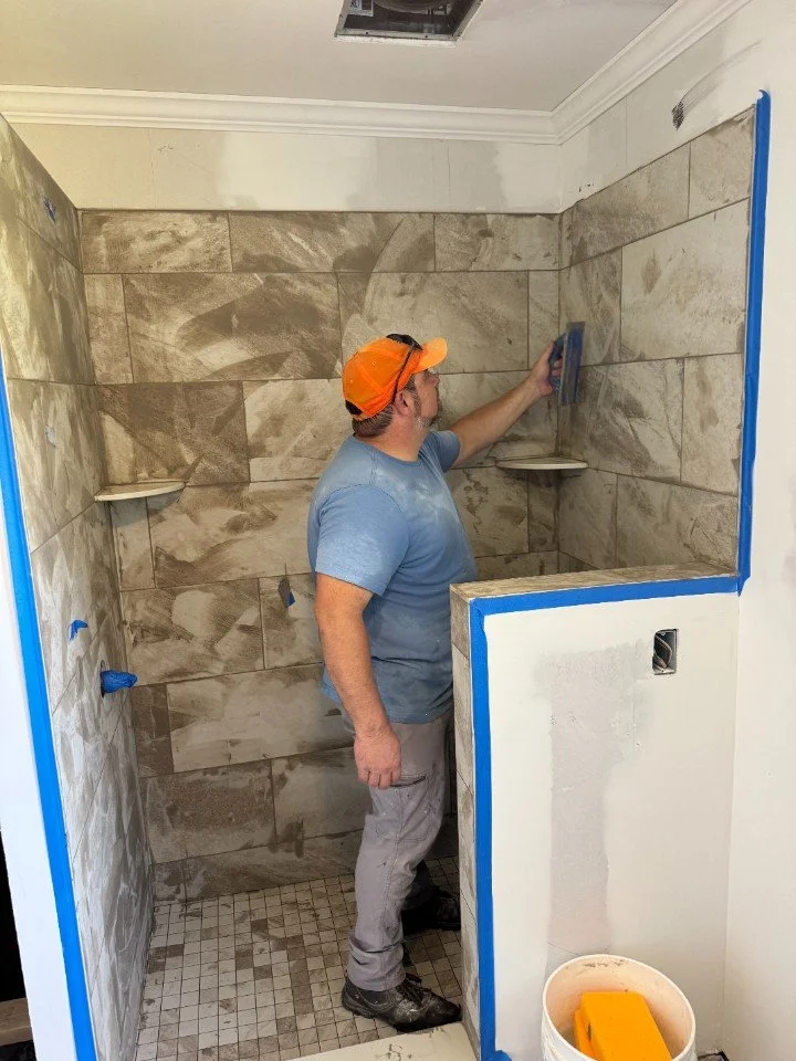 A man in a blue t-shirt, gray pants, and an orange cap installs large beige tile on a shower wall, using a trowel. The shower has a small built-in shelf, and the bathroom is under construction with blue painter's tape around the doorway and a bucket of tools in the foreground.