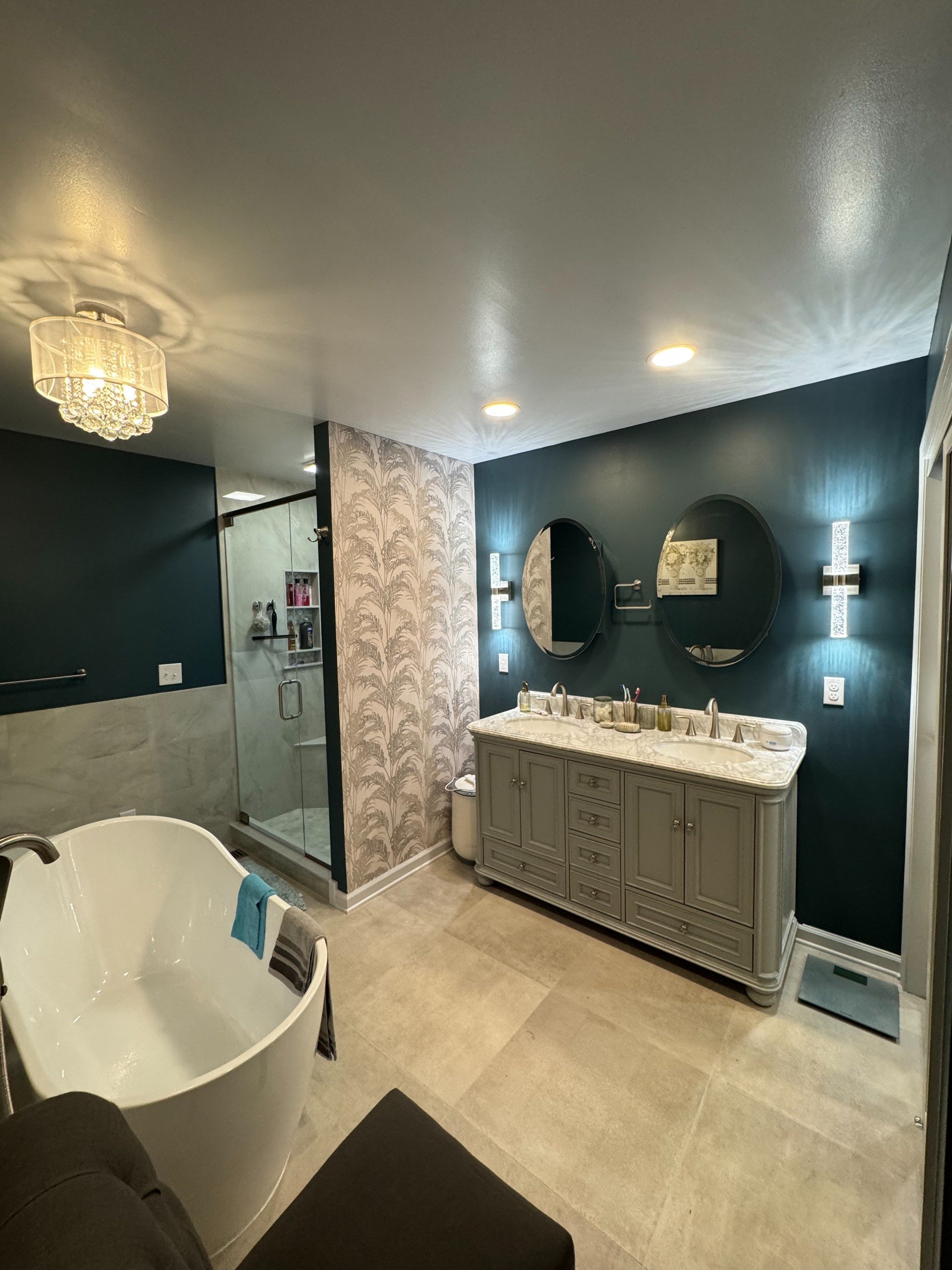 A modern bathroom with a freestanding bathtub, a vanity with two sinks, two mirrors, a glass-enclosed shower, and decorative lighting and wallpaper.
