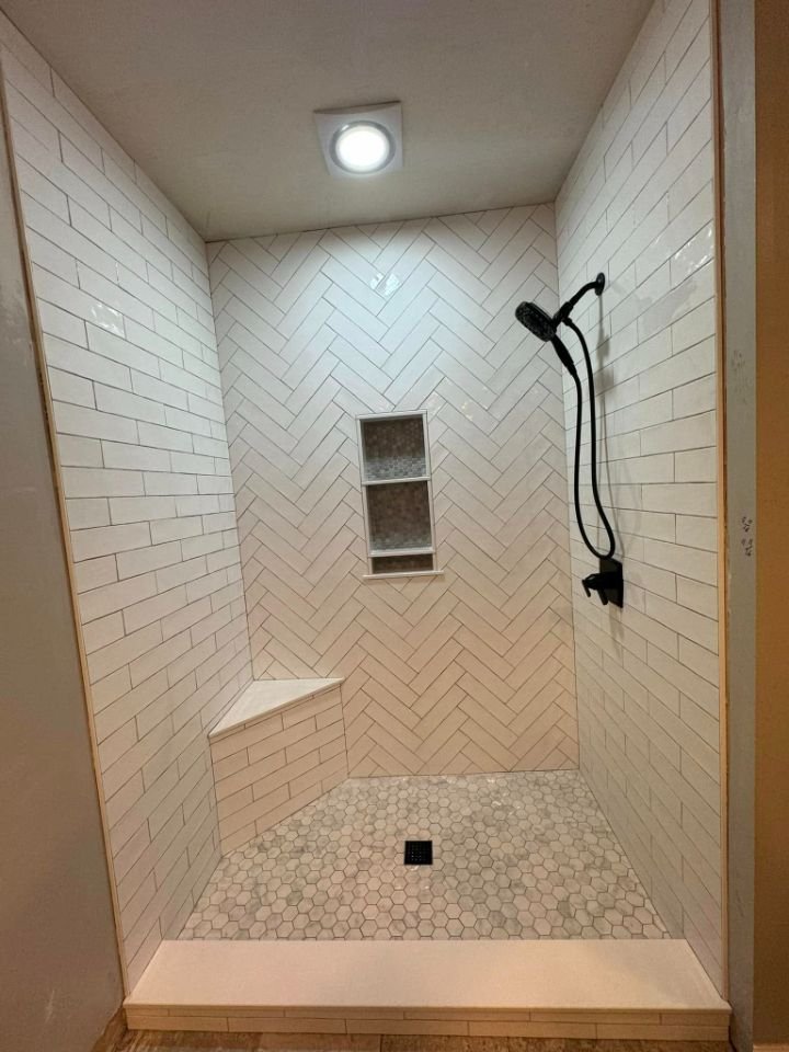 A modern walk-in shower with white subway tiles in a herringbone pattern on the walls, a built-in corner bench, a small recessed shelf, and hexagonal floor tiles. There is a black showerhead and a ceiling-mounted light fixture.