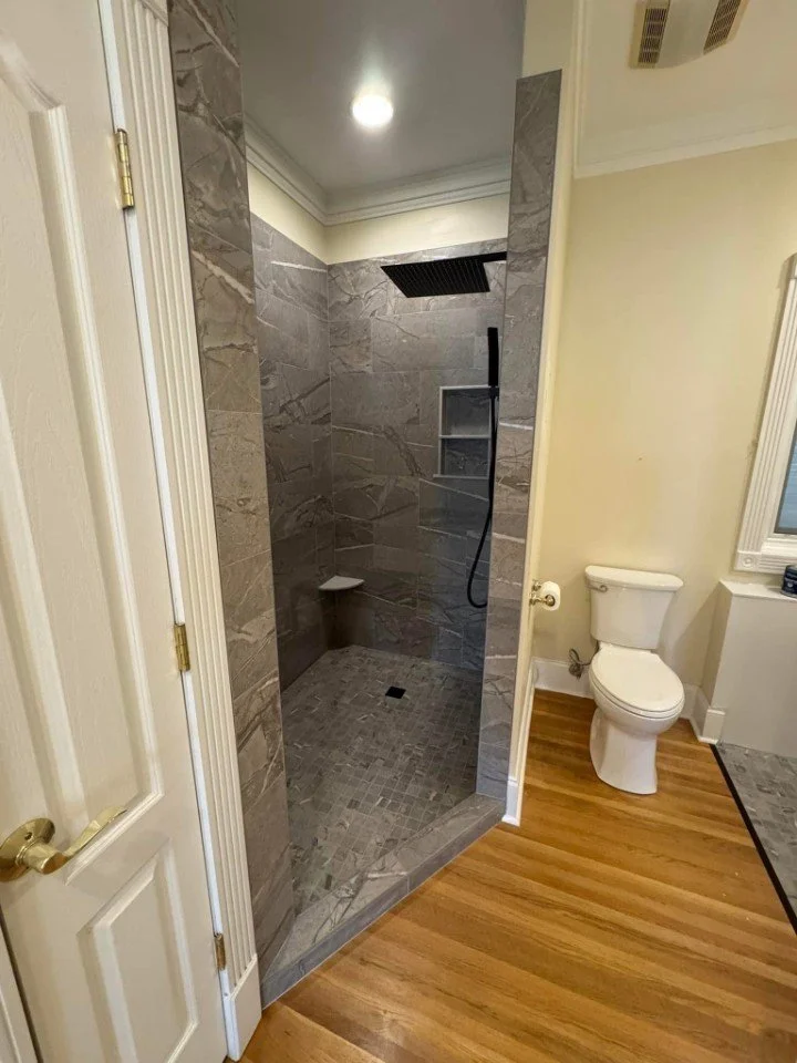 A bathroom with a glass-enclosed shower area featuring gray marble tiles, a built-in shelf, and a black showerhead. To the right, there is a white toilet next to a wall with a window, set on a wood floor.