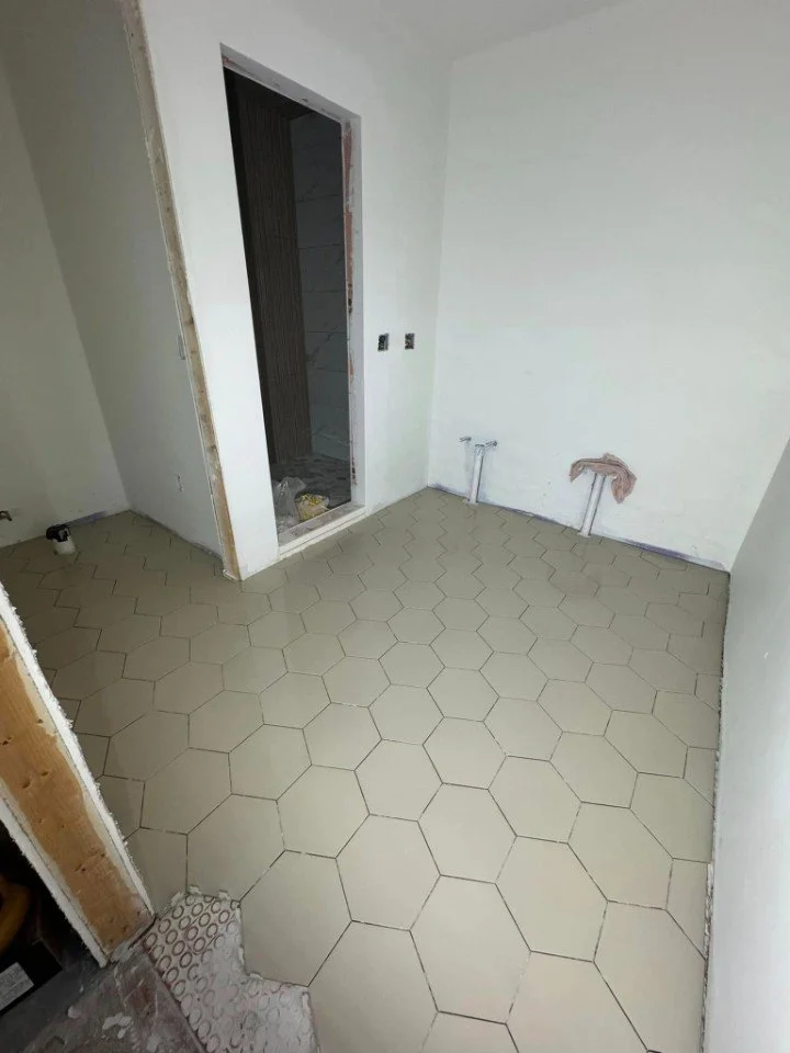 Unfinished room under construction with hexagonal beige tiles on the floor, white walls, and an open doorway leading to an adjacent room or bathroom.
