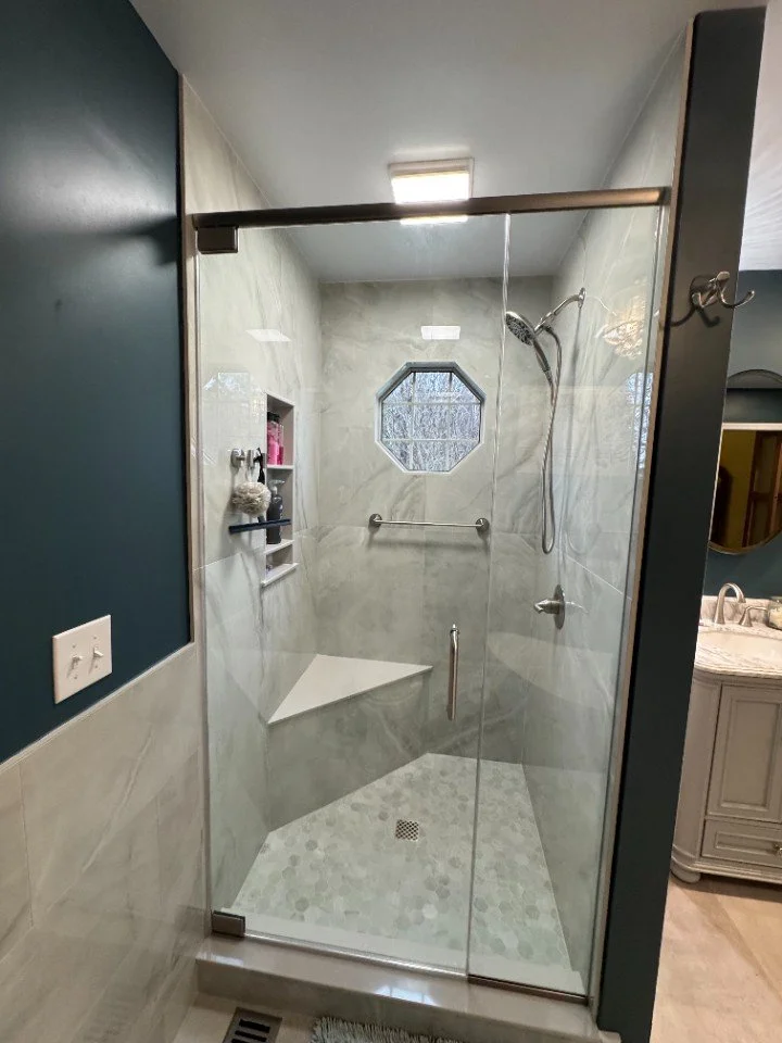 A modern shower with glass doors, beige marble walls, and a small octagonal window. Inside, there is a built-in nook with toiletries, a corner bench, a hand-held shower head, and a grab bar, with a floor drain and a sloped floor for water runoff.