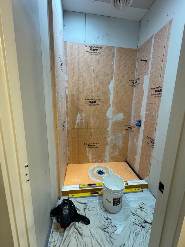 A small bathroom under renovation with unfinished walls covered in insulation material, a bucket on the floor, a level tool, a black handheld vacuum, and a cloth on the floor.