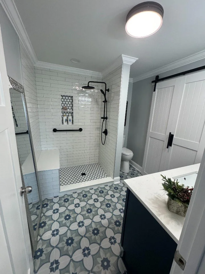 Bathroom with patterned tile floor, white subway tile shower with black fixtures, a built-in shelf in the shower, a toilet, and a navy blue cabinet with a white countertop holding a small plant. A sliding barn door is in the background, and a ceiling light is visible.