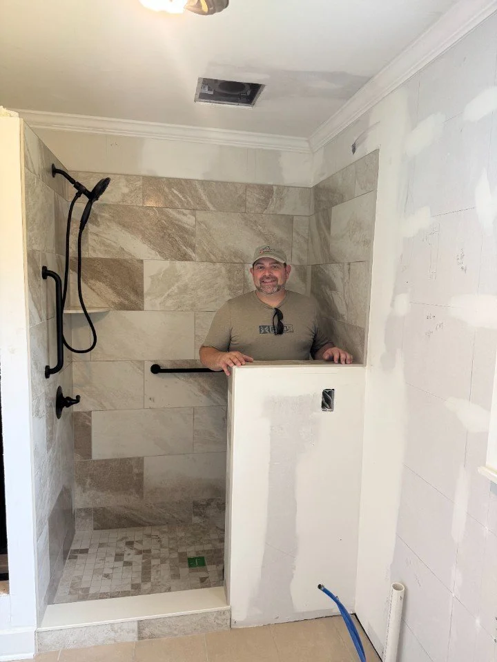 Man standing inside a partially built shower stall, smiling at the camera. The shower has tiled walls, a showerhead, and a grab bar. The enclosure is not yet finished, with an opening in the wall and construction materials visible.