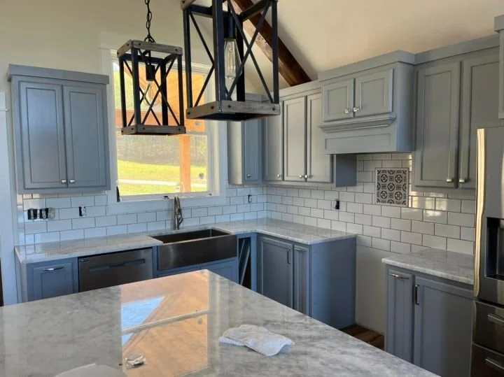 Light blue kitchen cabinets with white marble countertops, a farmhouse sink under a window, white subway tile backsplash, and black pendant lights hanging above the island in a modern kitchen.