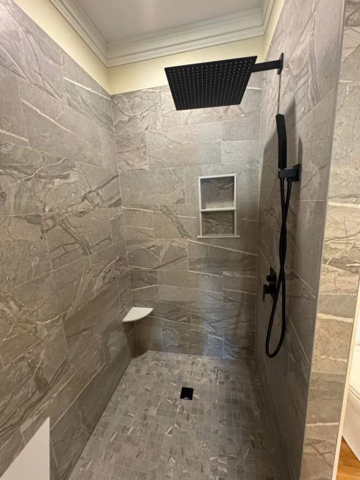 A modern walk-in shower with large beige marble walls, a black overhead rainfall showerhead, and a separate handheld showerhead with a black hose. There are two small niches built into the wall for toiletries and a small corner shelf.