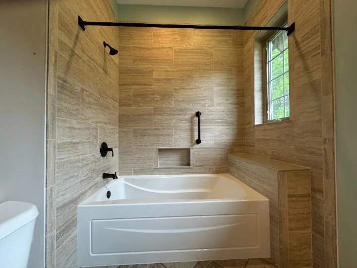 Bathroom with a bathtub surrounded by beige tiled walls, black fixtures, and a small window with a view of greenery outside.