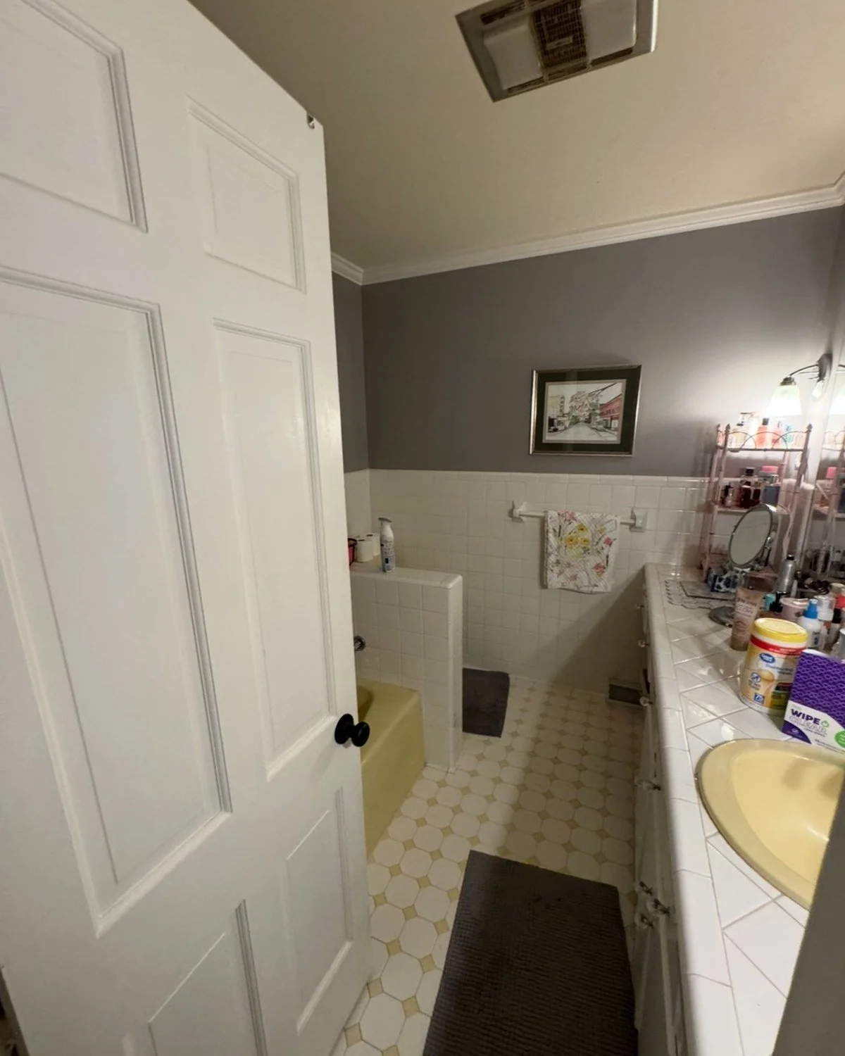 Bathroom with yellow sink, white tiled counter, gray wall, framed picture, towel rack, small bathtub, various toiletries, and a mirror.