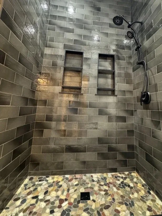 A shower with dark gray tile walls, a pebble tile floor, and two built-in shelves. It has a black showerhead and fixtures.