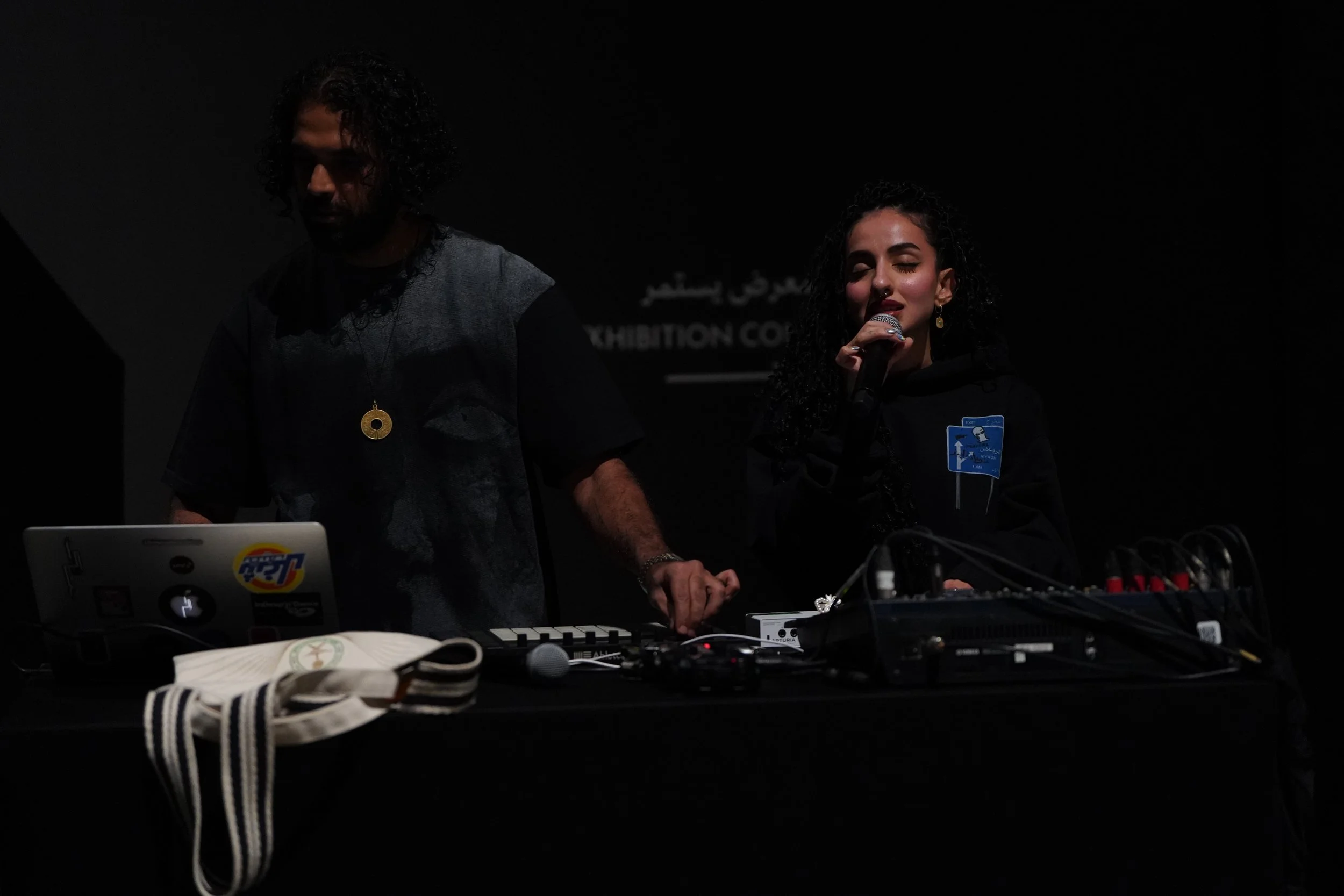 Two people, a man and a woman, performing music with electronic equipment on a dark stage. The woman is holding a microphone and appears to be singing, while the man is operating a keyboard or synthesizer. The setup includes laptops, a mixer, and various cables, with a face mask and some bags visible in the foreground.