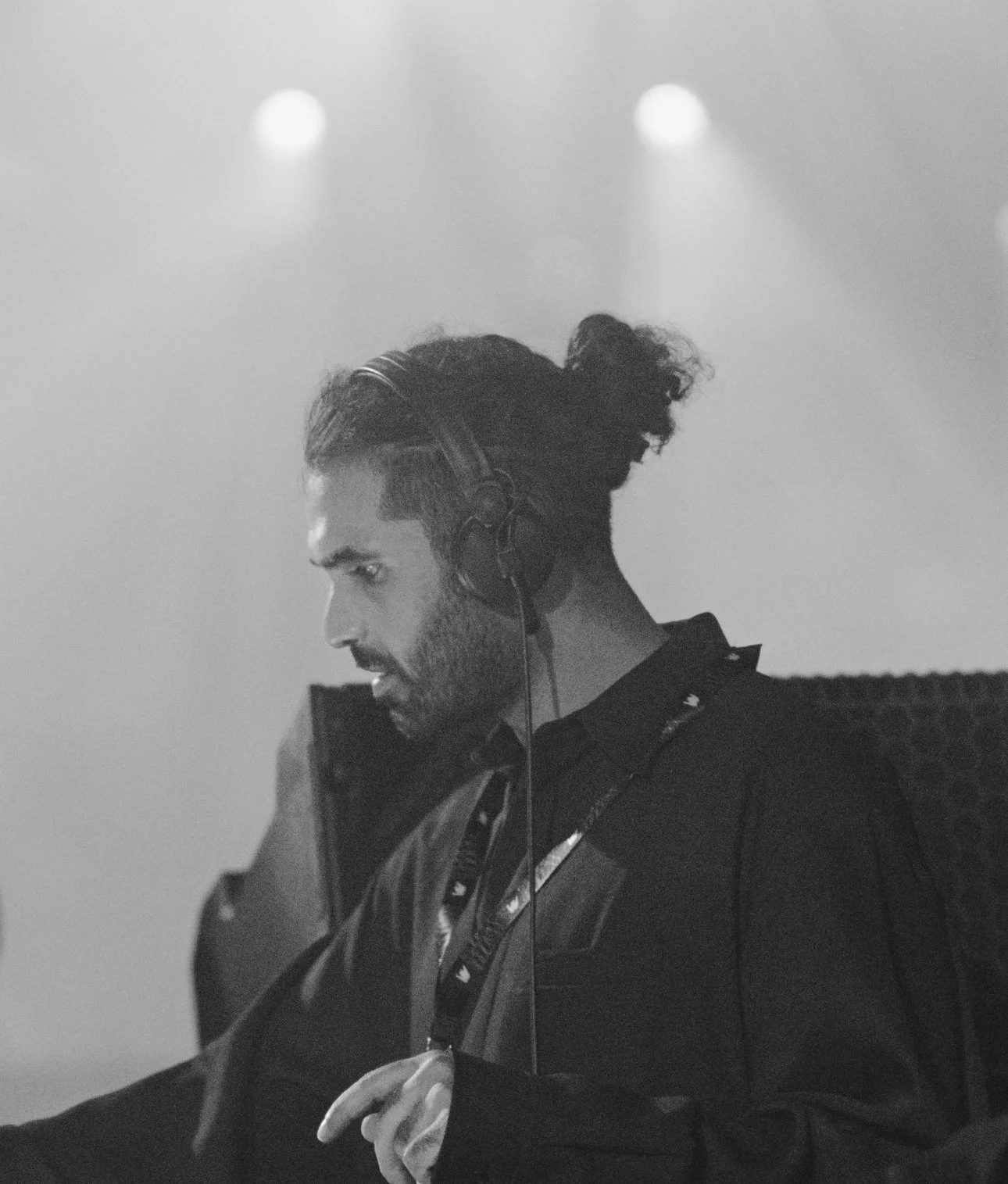 Black and white photo of a man with long hair tied in a bun, wearing headphones and working on a DJ mixer, focused on his equipment.