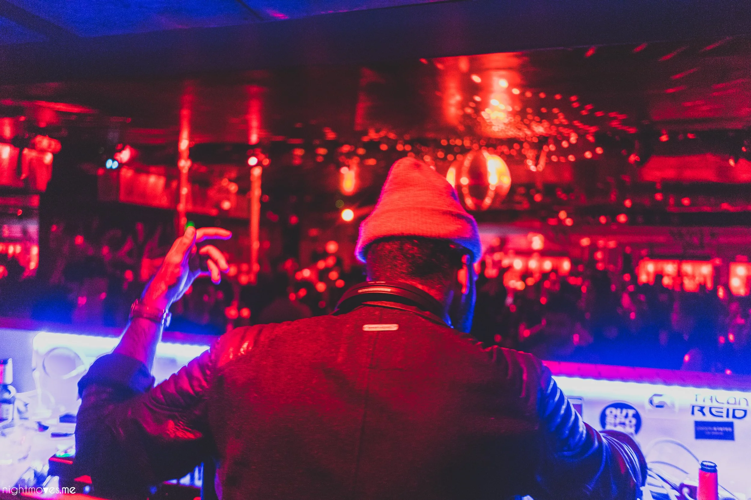 A person wearing a beanie and jacket is standing in a nightclub or bar with red and purple lighting, facing away from the camera, surrounded by other people and illuminated by colorful lights.