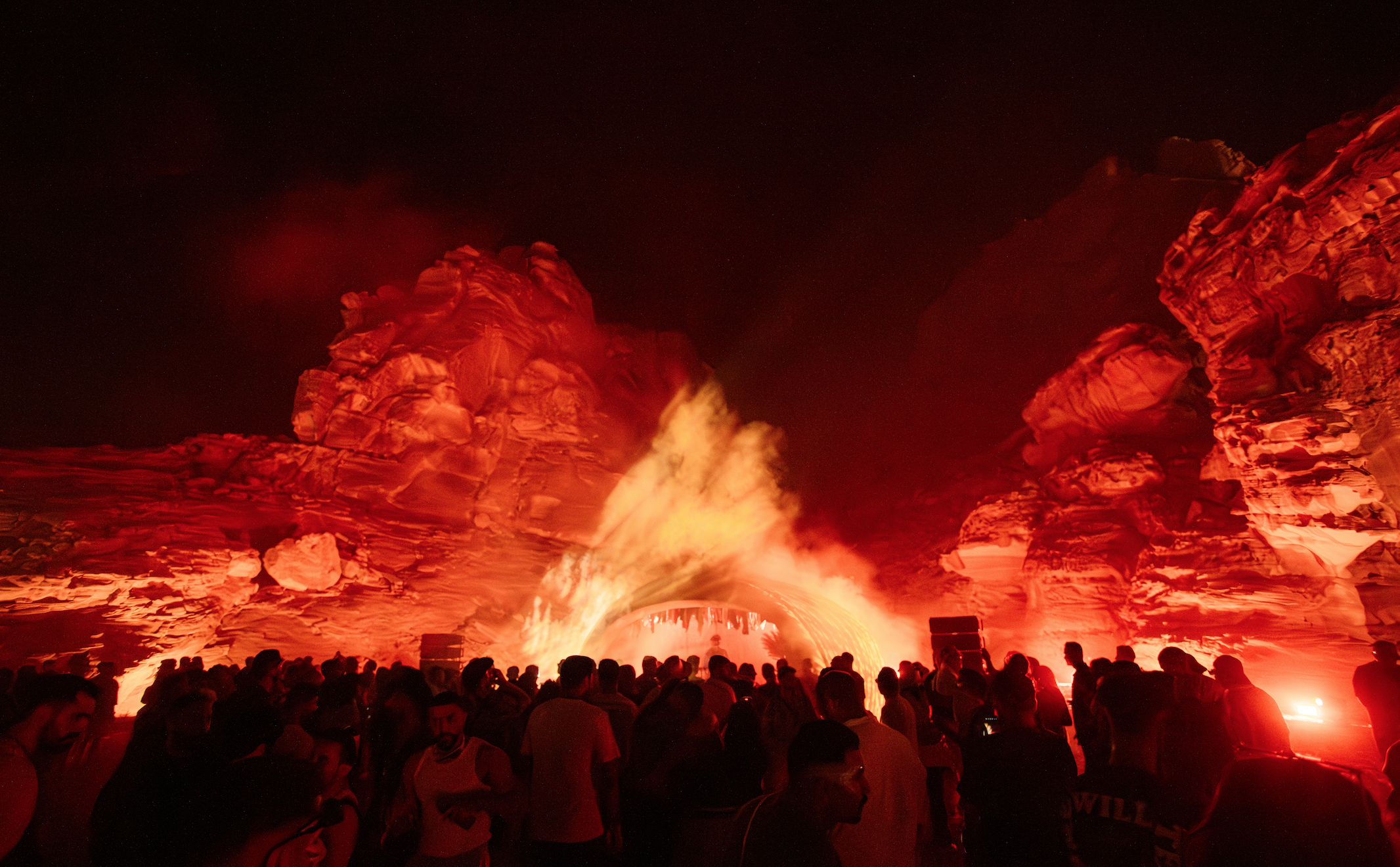 A large outdoor concert or event taking place at night with a crowd of people watching a fiery spectacle of flames and pyrotechnics against rock formations, illuminated in red and orange.