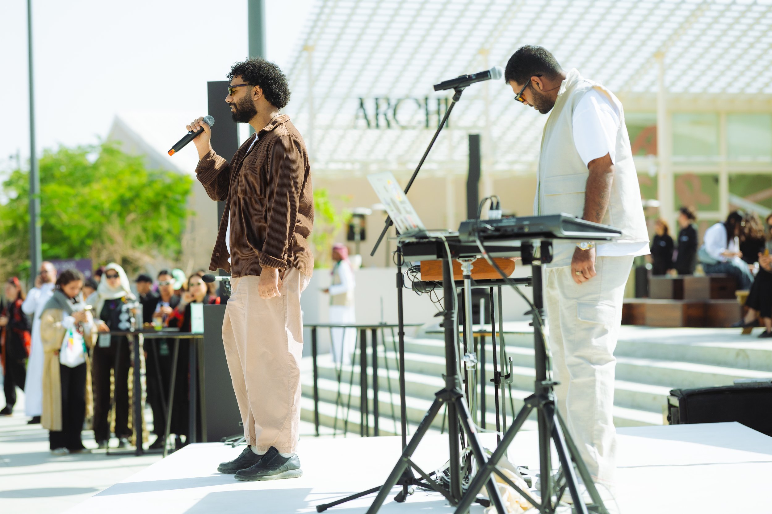 Two men, one holding a microphone, are on a stage at an outdoor event with a crowd in the background. One man is dressed in a brown jacket and beige pants, the other in a white shirt and cream-colored pants. There is audio equipment and a microphone stand on the stage.