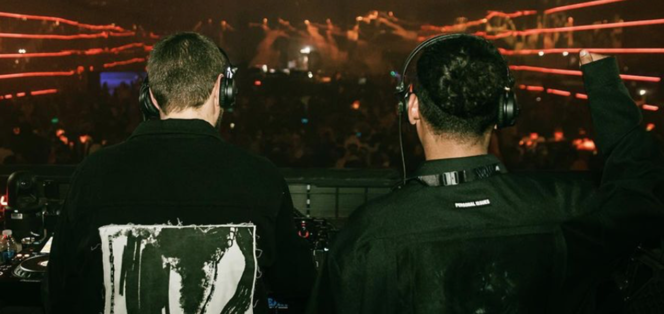 Two DJs wearing headphones performing at a concert with orange laser lights and a crowd in the background.
