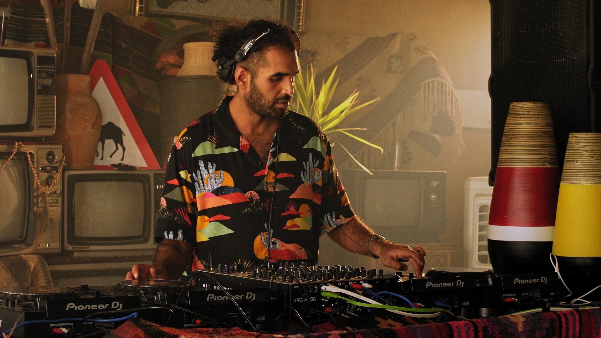 A DJ wearing a rainbow-colored tropical shirt and headphones playing music behind a Pioneer DJ mixer in a retro-themed setting with vintage televisions, a warning sign, large decorative vases, and stacked colored vinyl records.