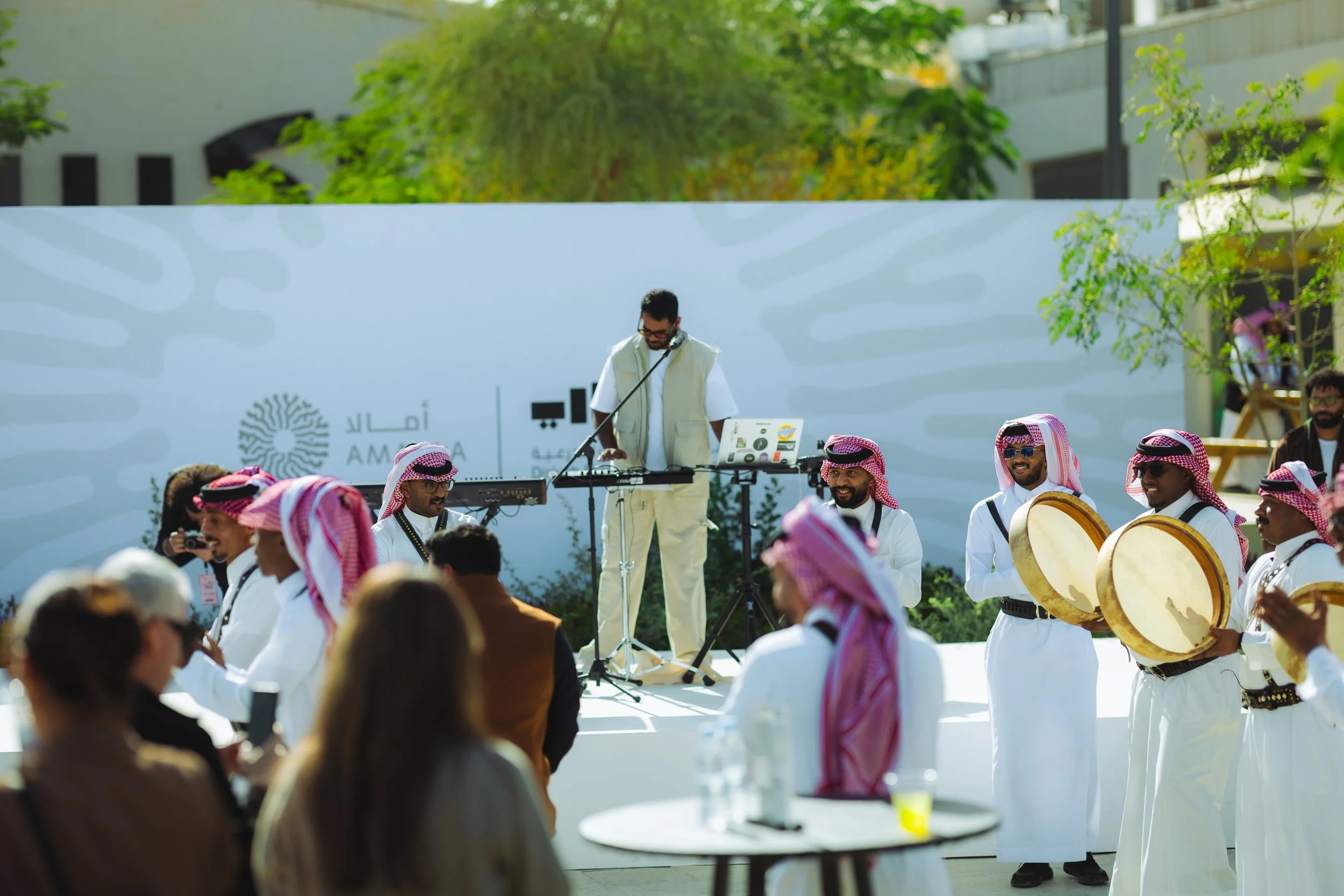 Outdoor event with musicians in traditional Middle Eastern attire, including white robes and red-and-white-checkered headscarves. One person is standing at a DJ setup, wearing glasses and a white vest, while others are playing drums. Audience members are in the foreground, some taking pictures. The background features a large white wall with minimal text and trees, indicating a sunny day.