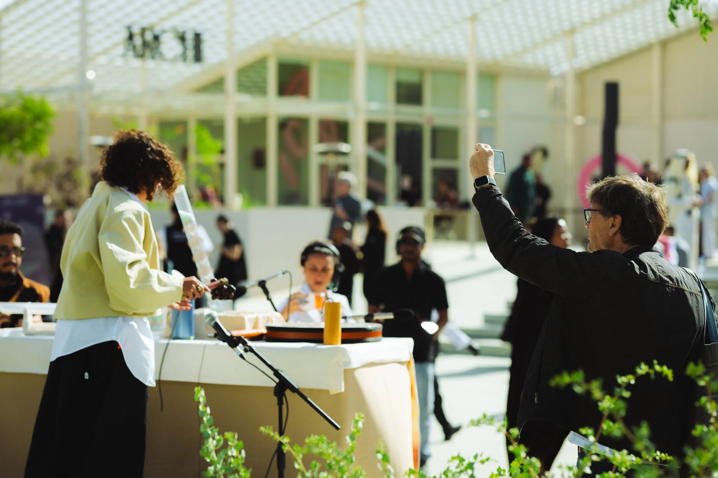 People at an outdoor social event, with one person taking a photo and others seated at tables, in front of a modern building with glass and a grid-like roof.