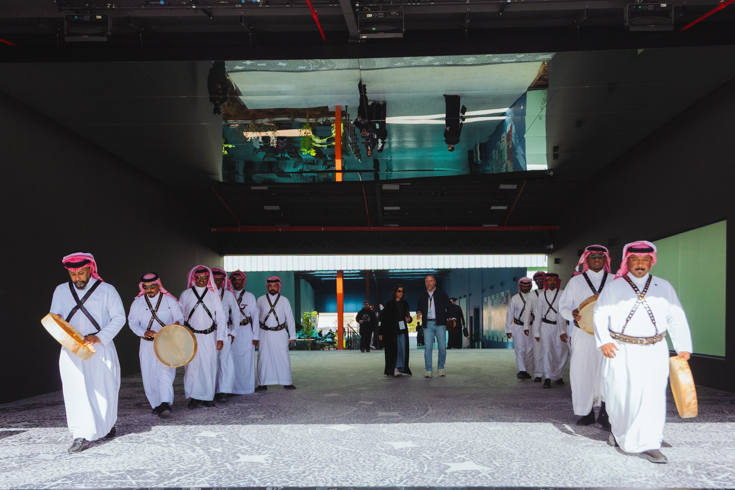 Group of men dressed in traditional Middle Eastern attire playing drums and tambourines during an event in a modern building with reflective ceilings.