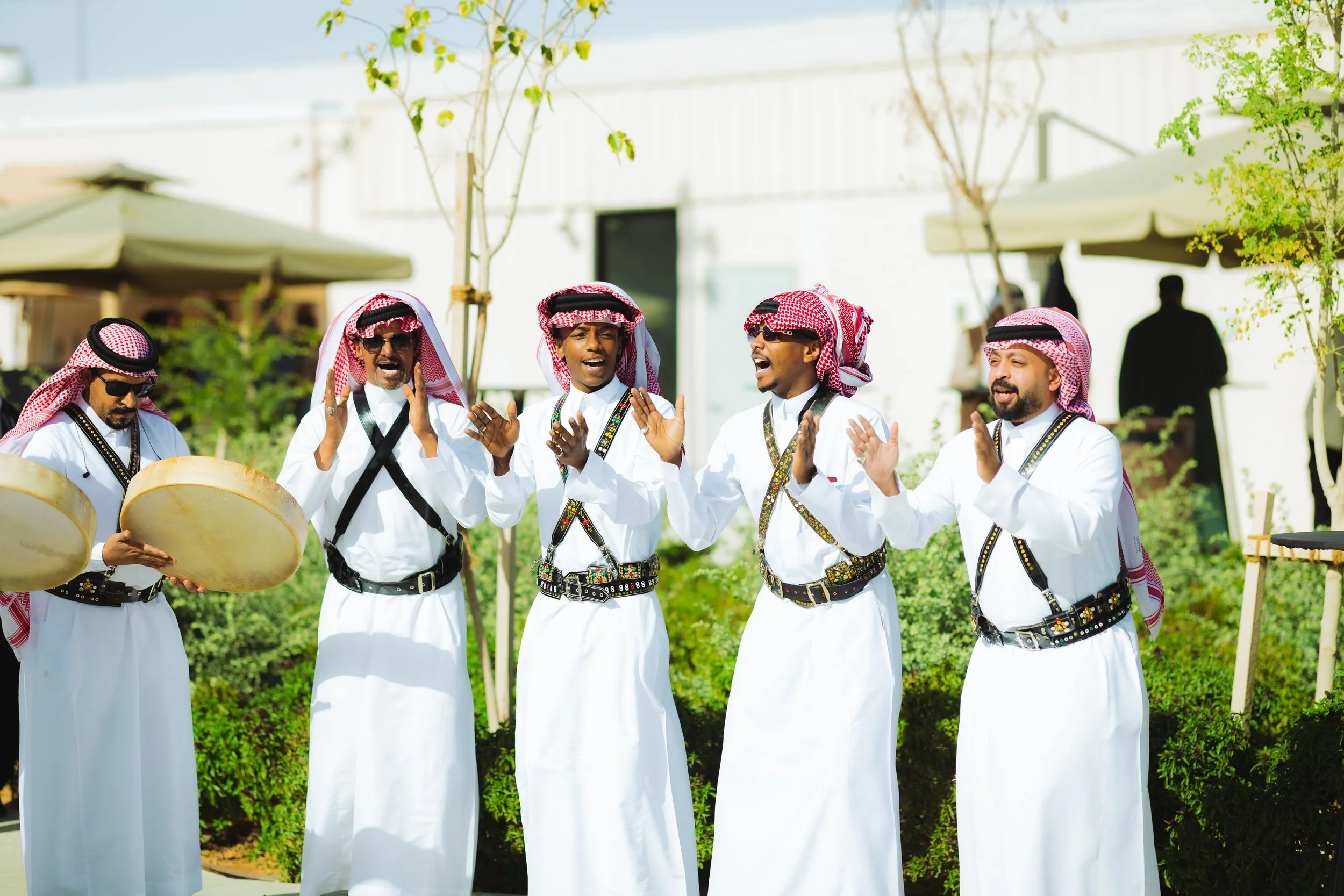 A group of five men dressed in traditional Middle Eastern attire, including white thobes, red and white checkered headscarves, and sunglasses, are clapping and singing outdoors during daytime. One man is playing a tambourine. They are surrounded by greenery and outdoor furniture with umbrellas.