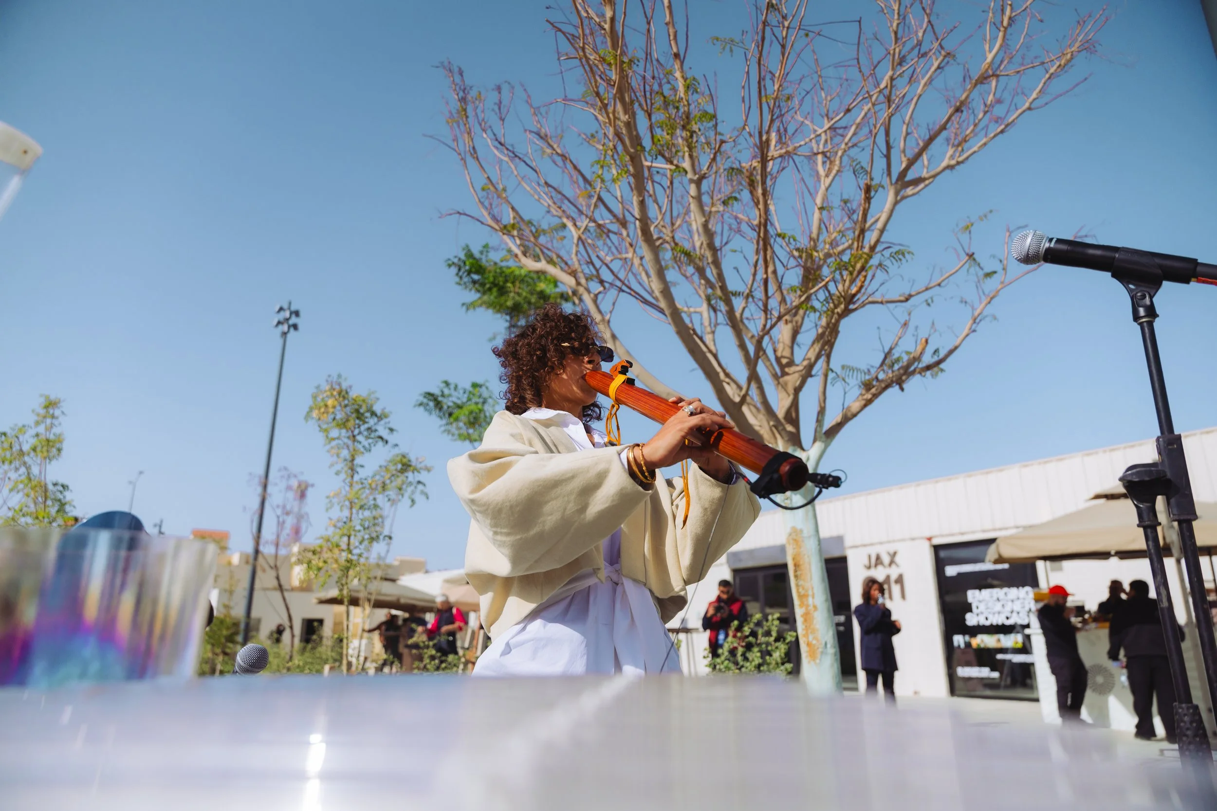 A person with curly hair and sunglasses playing a wind instrument outdoors during daytime, with a clear blue sky, leafless tree, and people in the background.