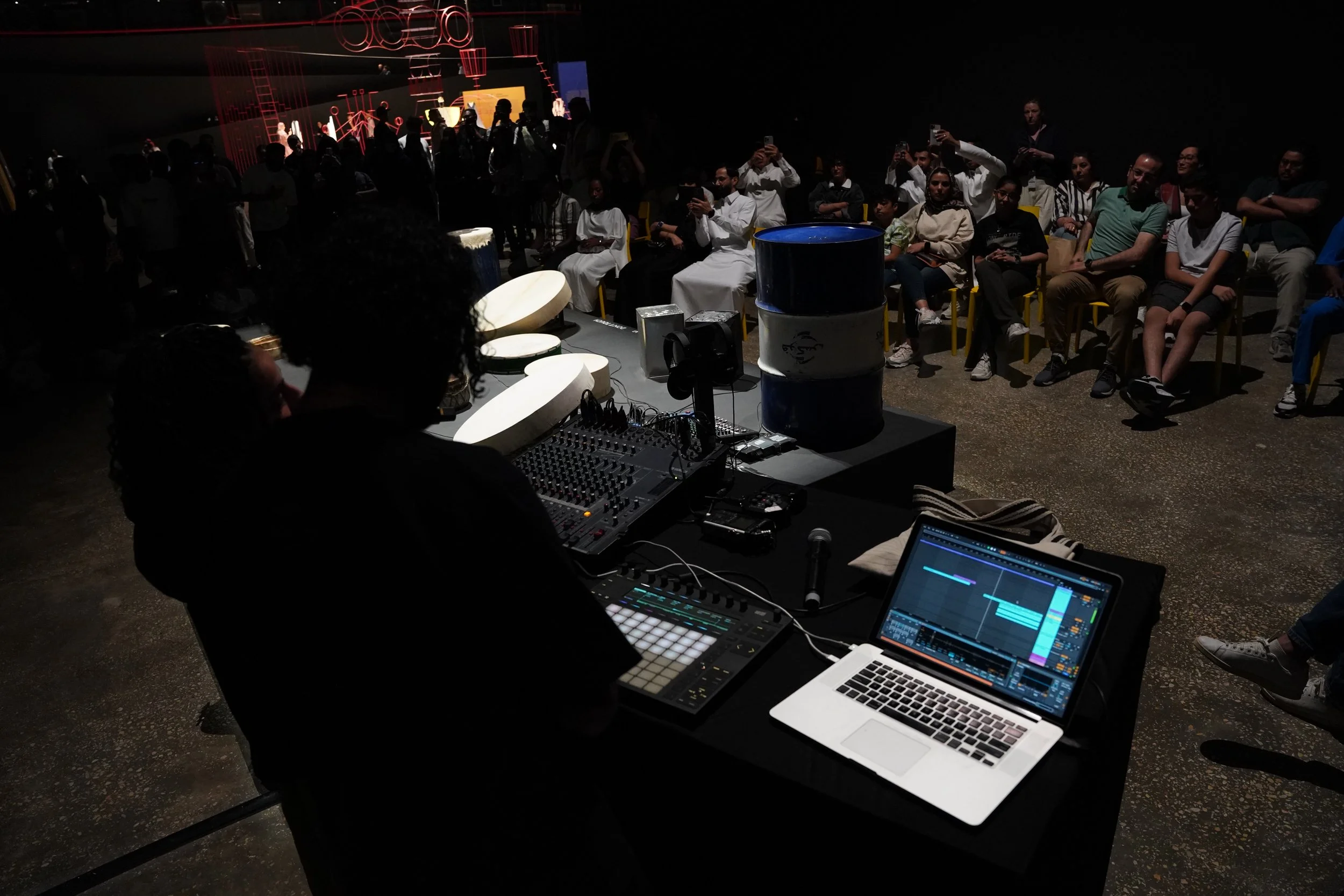Audience seated in a dark room watching a performance or presentation, with some people taking photos. A DJ or music producer's equipment, including a laptop and mixing console, is visible in the foreground.