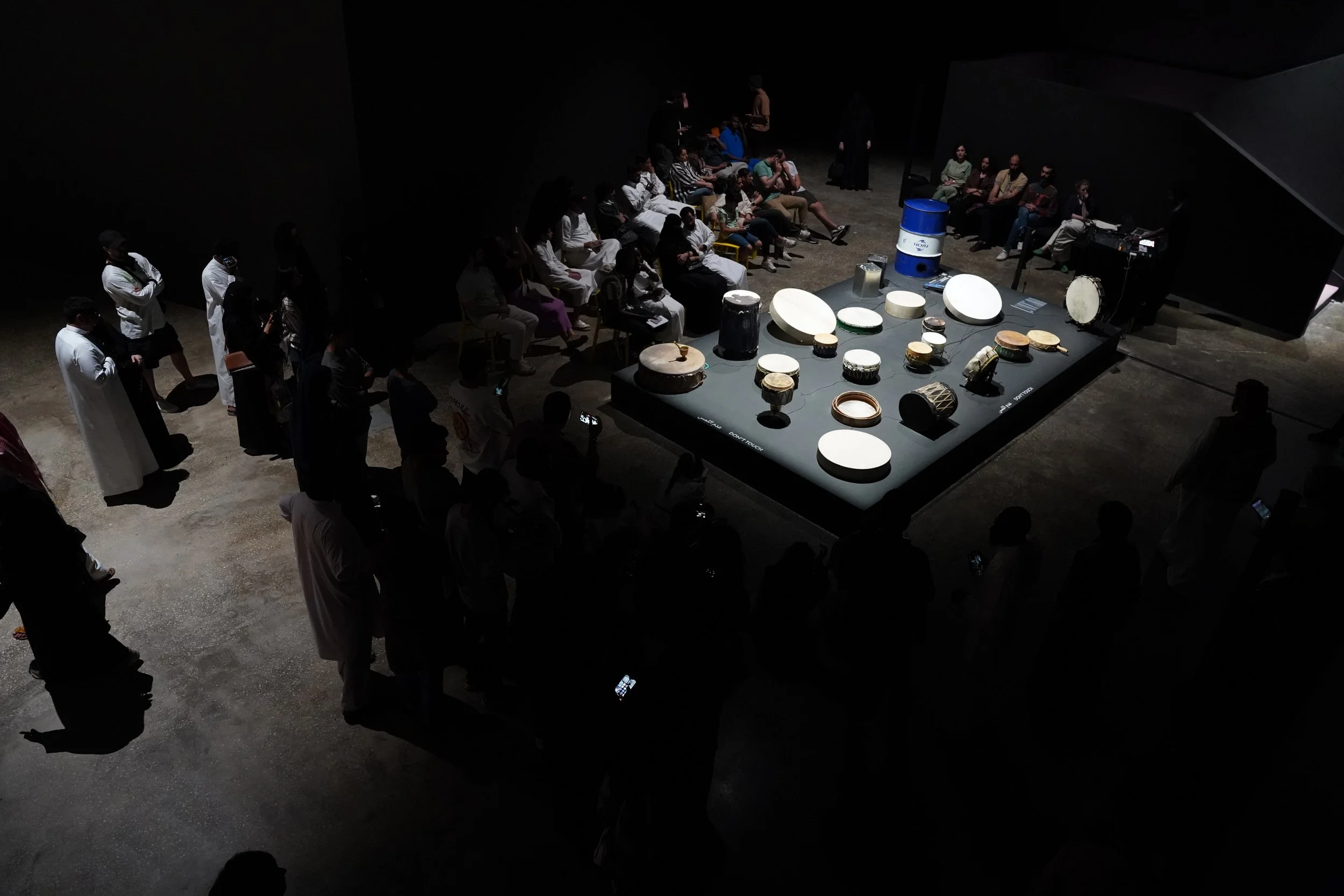 An indoor concert or event with a large group of people seated in a room, facing a stage area with various drums and musical instruments, some individuals standing and watching.