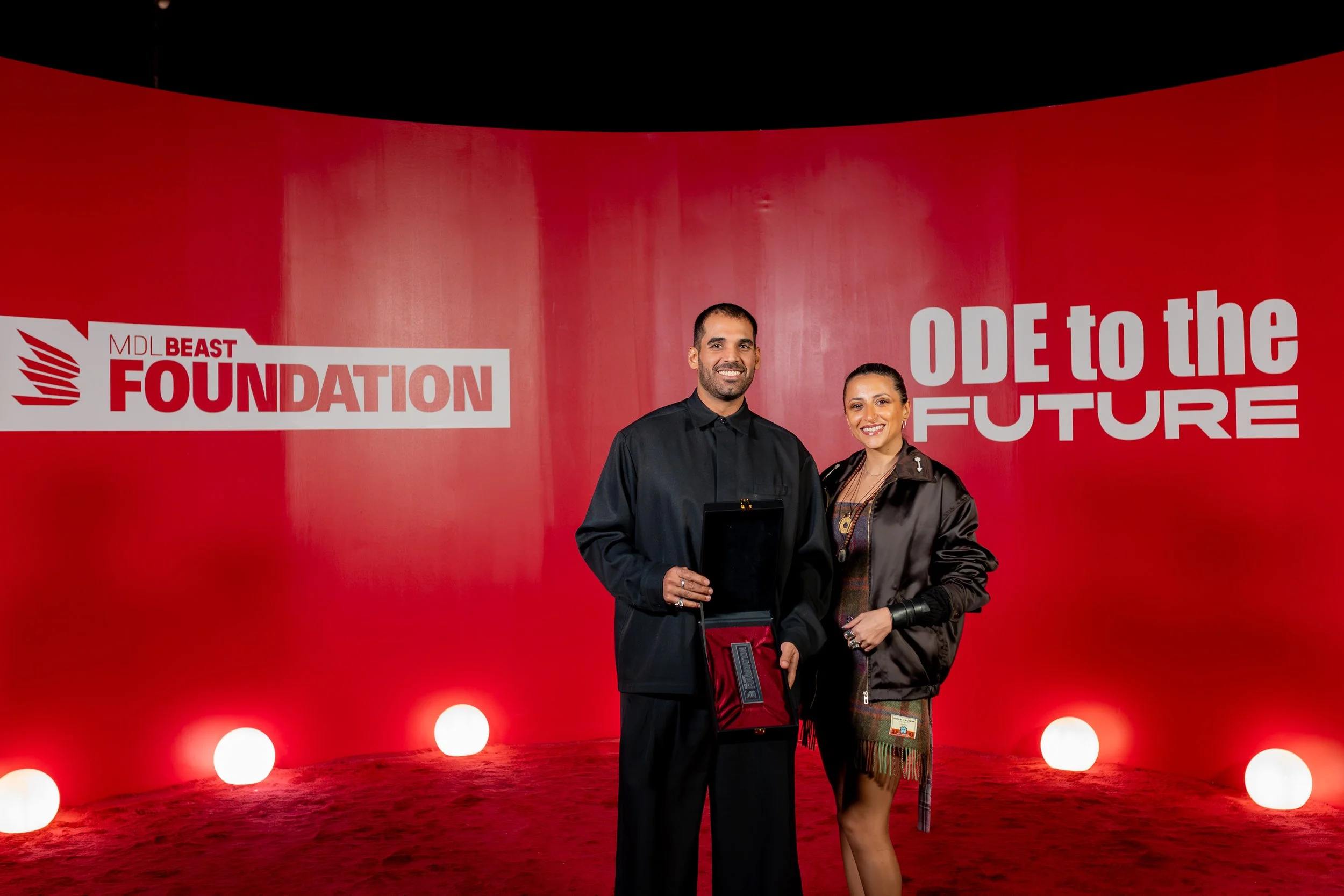 Two people standing in front of a red background with white text and circular lights at the bottom; one man holding an award and a woman smiling.