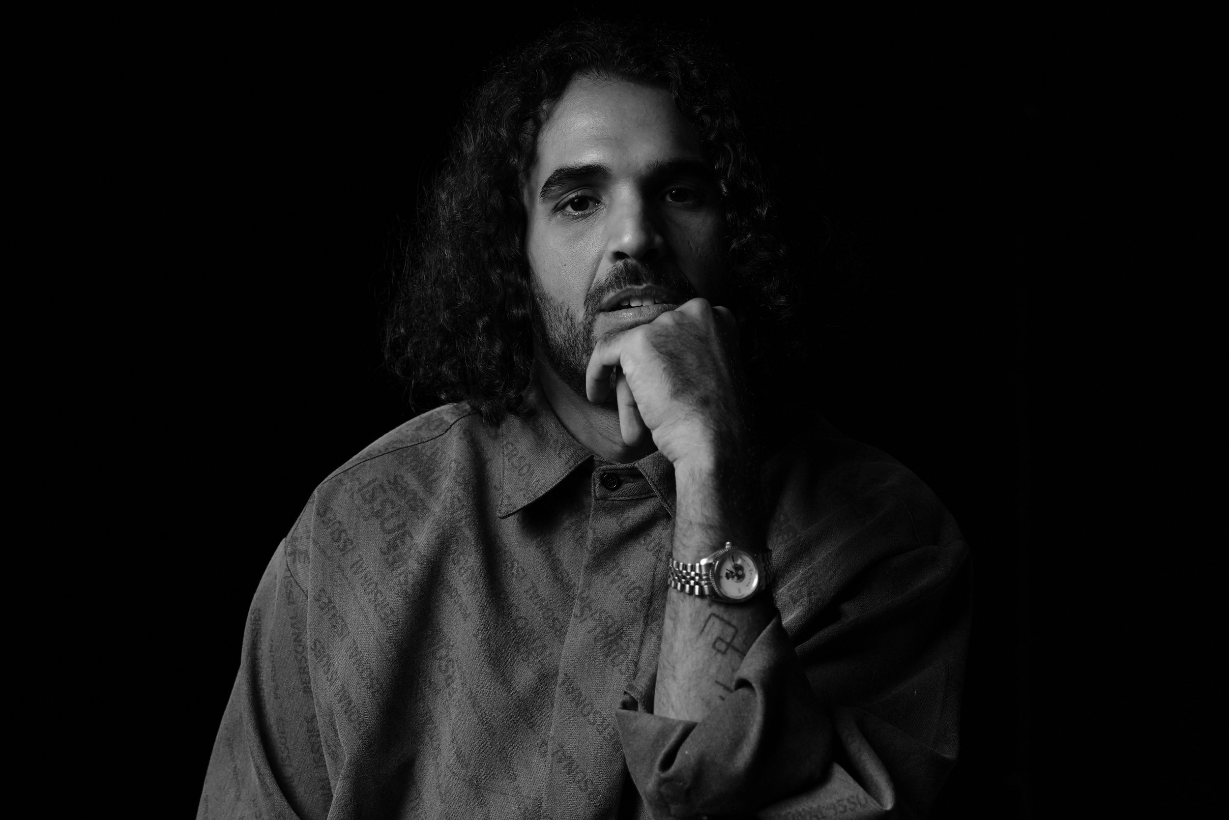 Black and white portrait of a man with curly hair and a beard, wearing a collared shirt and a watch, resting his chin on his hand against a dark background.